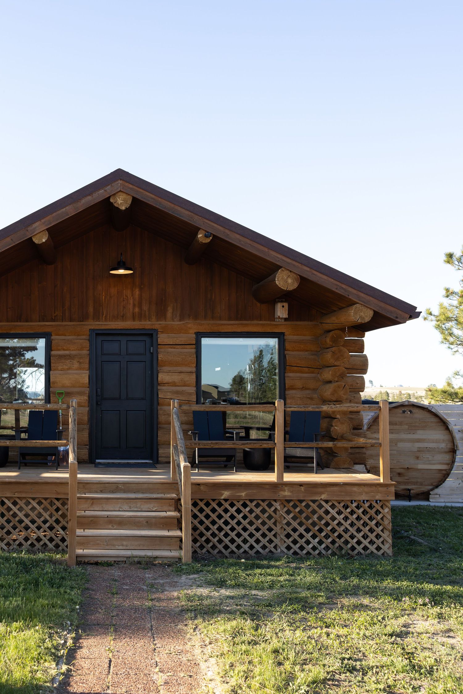 Rustic log cabin with wooden porch, black front door, large windows, and a barrel hot tub on a sunny day.