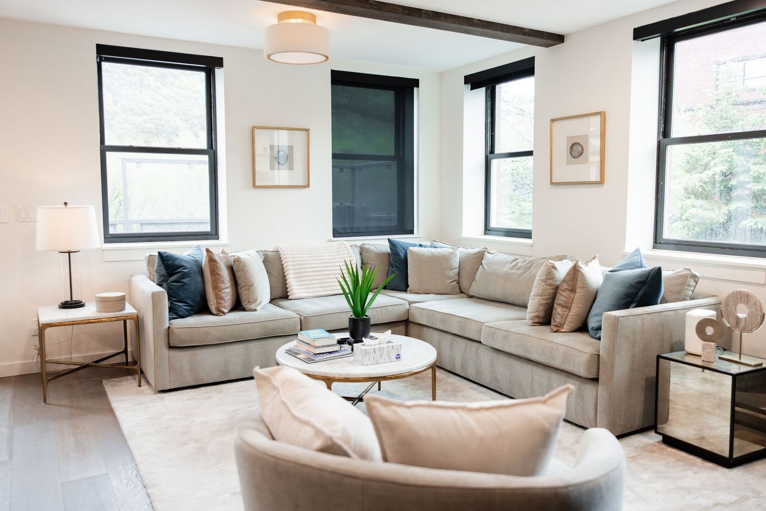 Modern living room with gray sectional sofa, navy accent pillows, white round coffee table, and large windows with black frames.