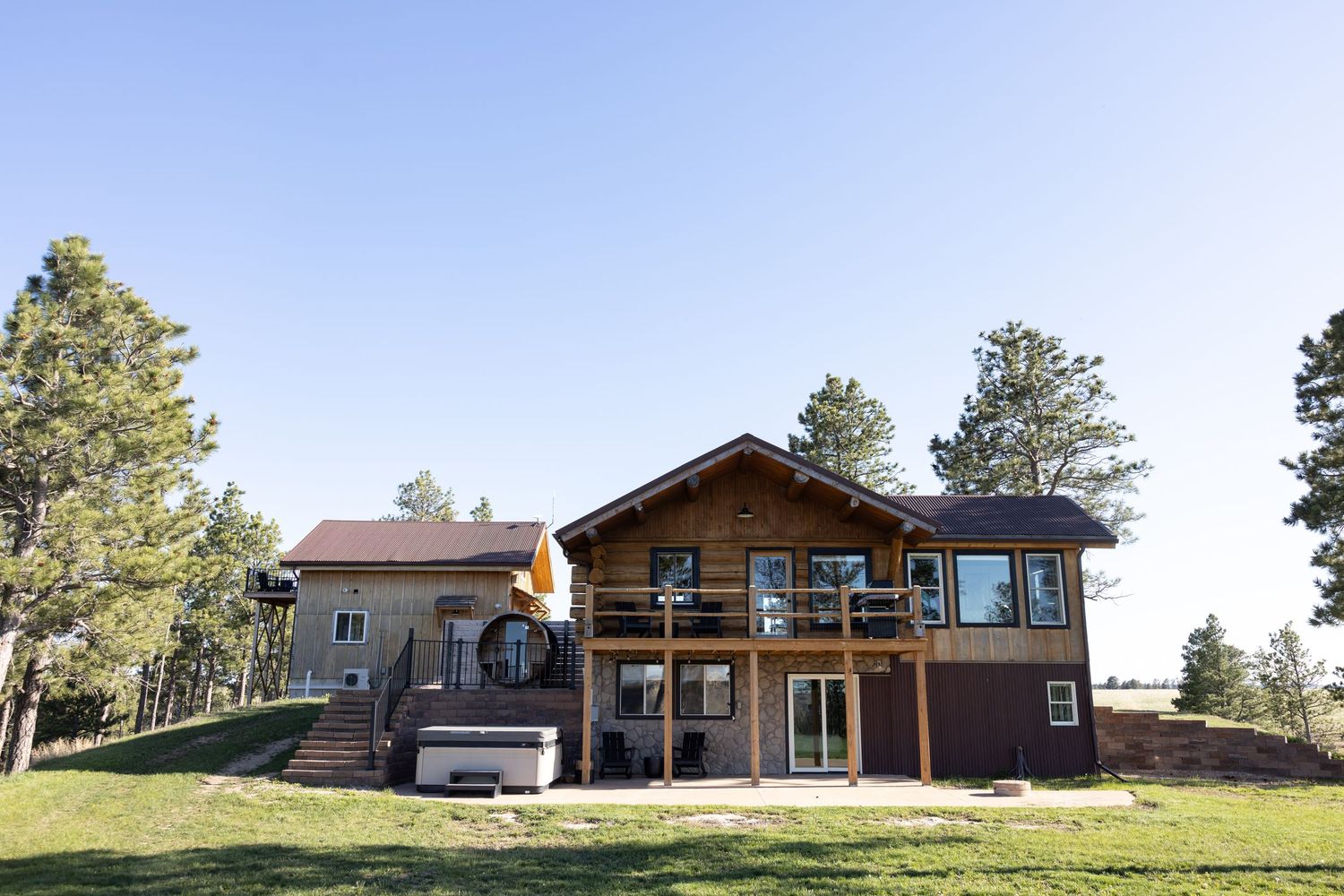 Large two-story log cabin home with wrap-around deck surrounded by pine trees on a green hillside.