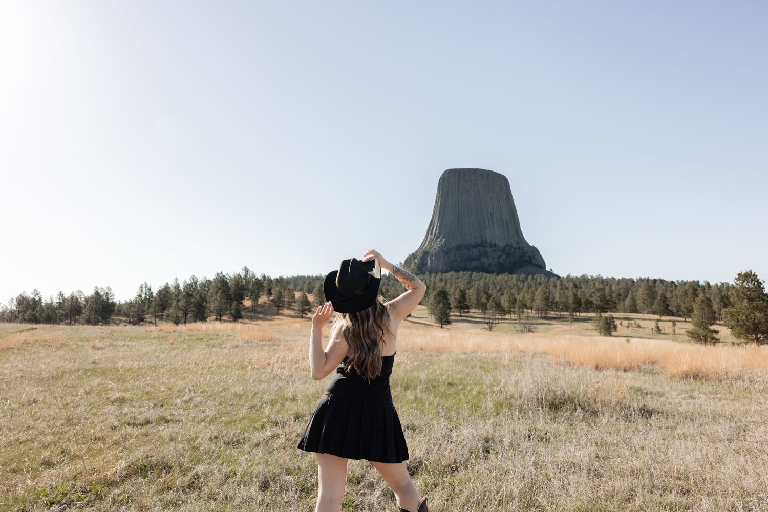 Woman in black dress and cowboy hat stands before Devils Tower National Monument in Wyoming on a clear day.