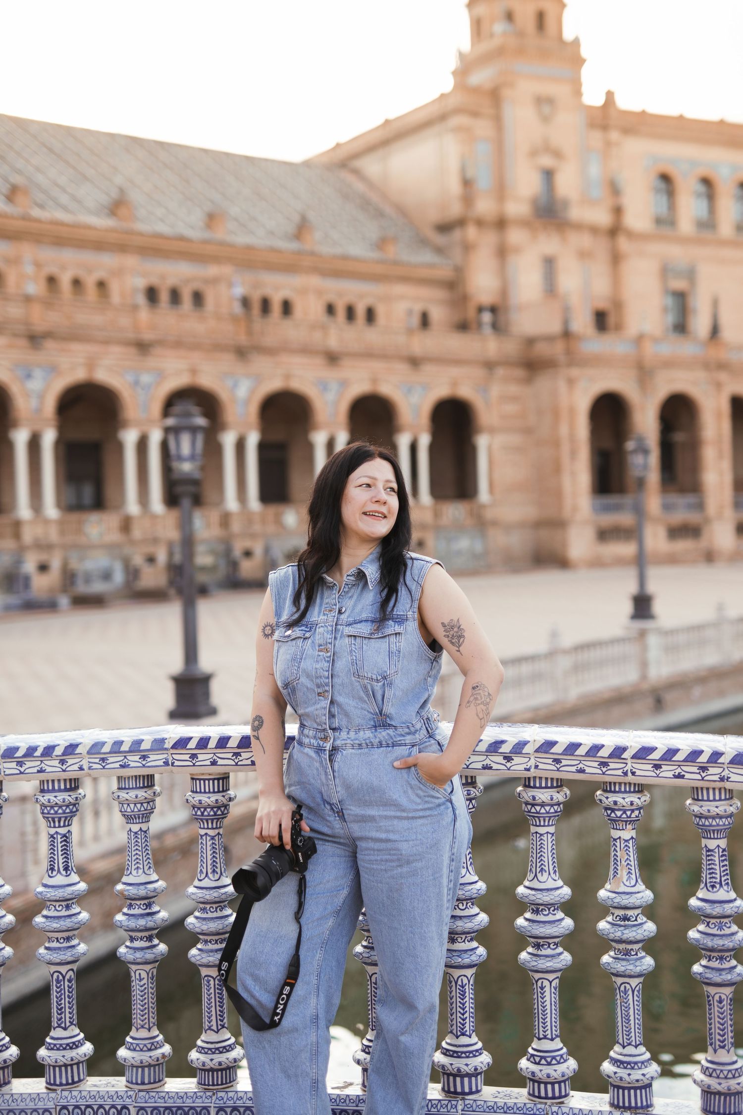 Woman in denim jumpsuit with tattoos poses at Plaza de España, Seville, holding a camera by ornate tiled railing.