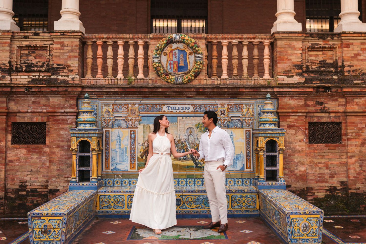 Couple in white attire posing at Plaza de España Seville, surrounded by colorful ceramic tilework and brick architecture.