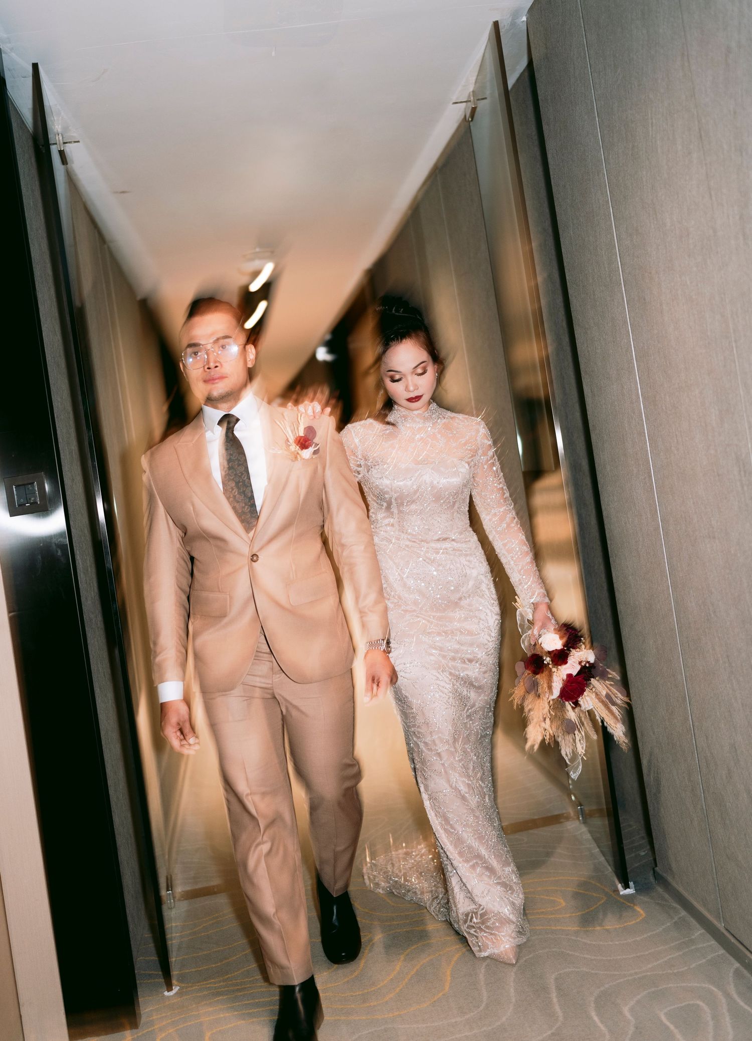A couple walking down a hotel hallway, with the bride in a lace gown holding a bouquet with groom for malay wedding at Novotel on Stevens.