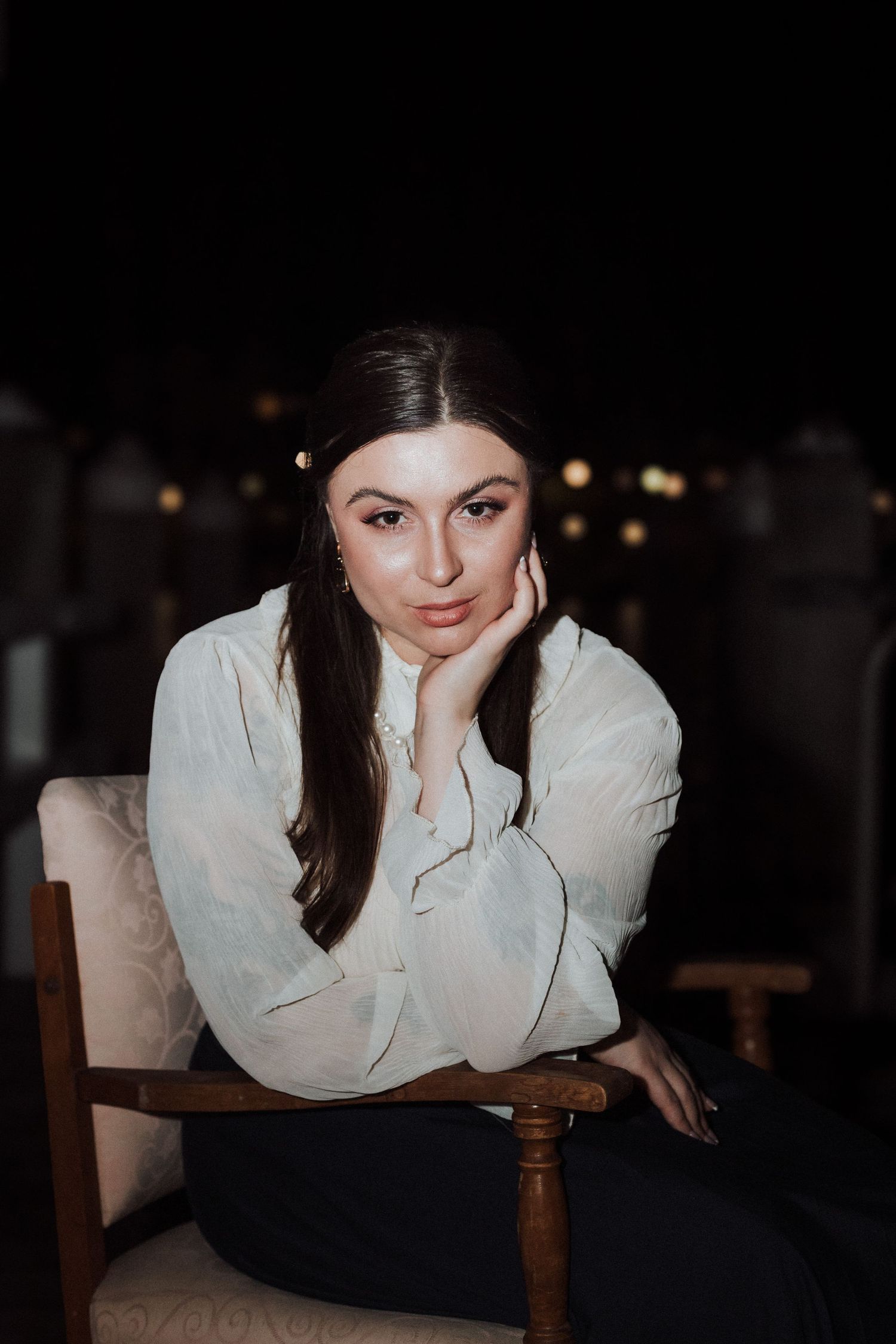 A moody portrait of a woman in white blouse sitting in wooden chair with ambient lighting.