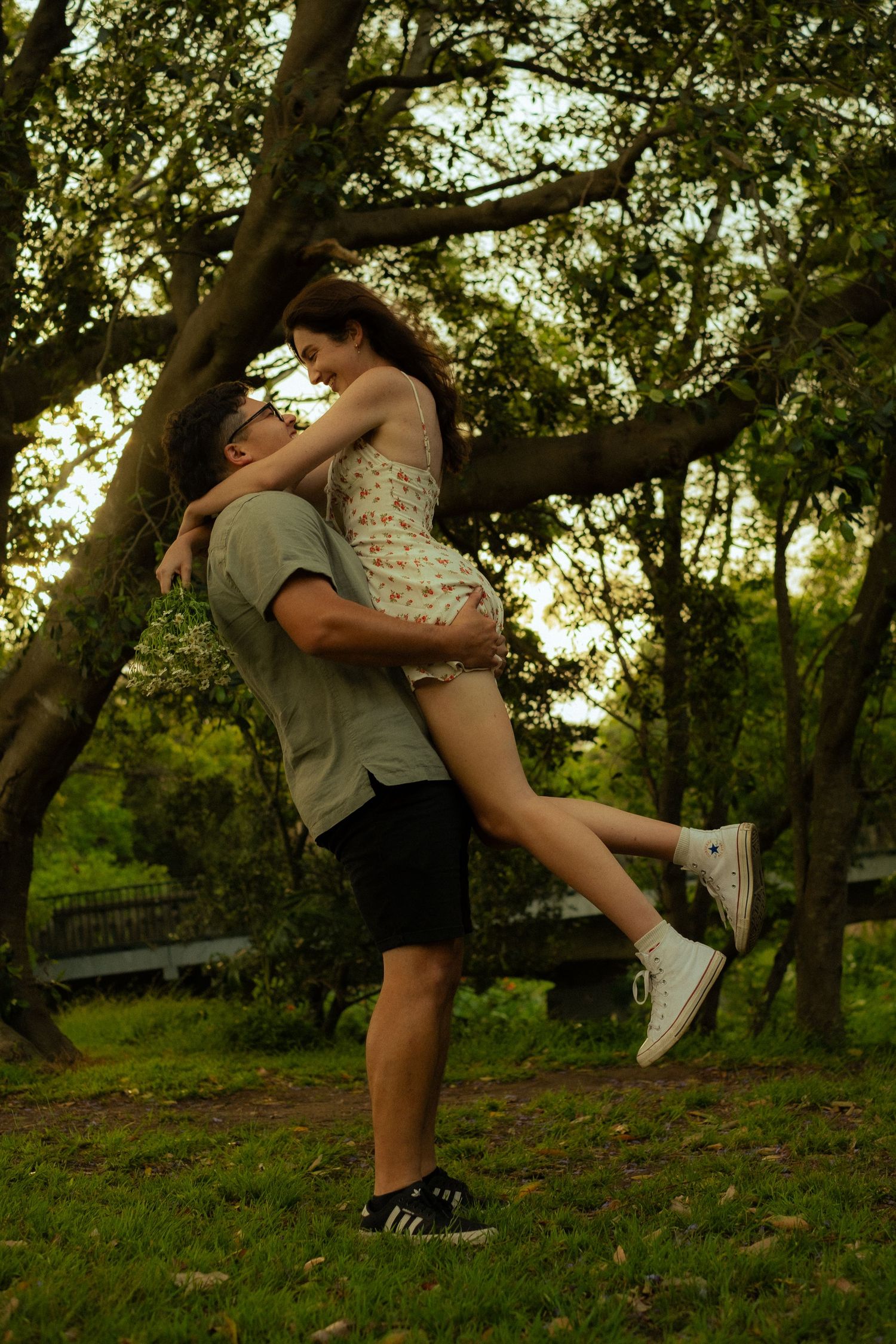 Playful couple embracing in park during golden hour with trees in background.
