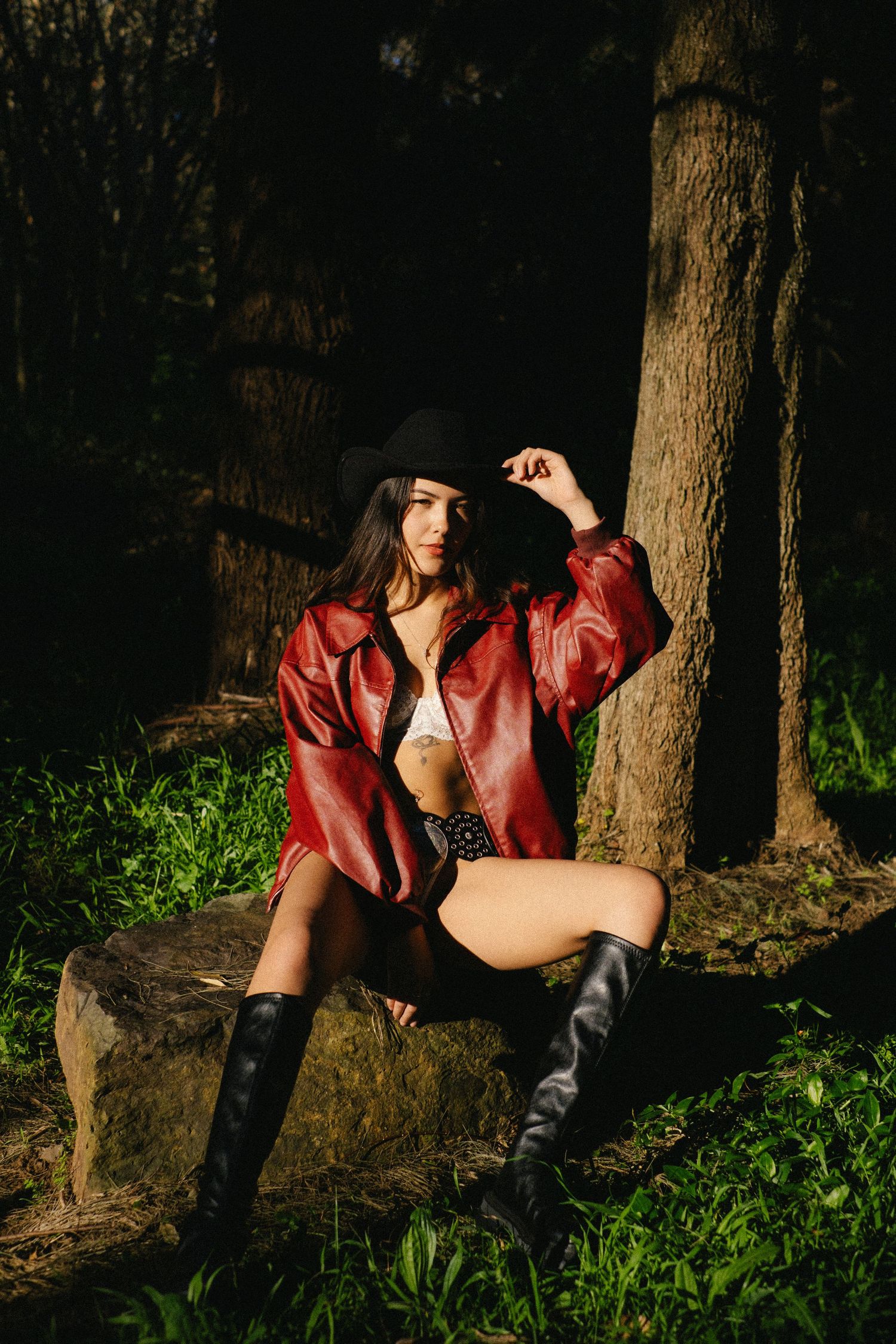 Model in red leather jacket and black boots poses against tree trunk in moody forest setting.
