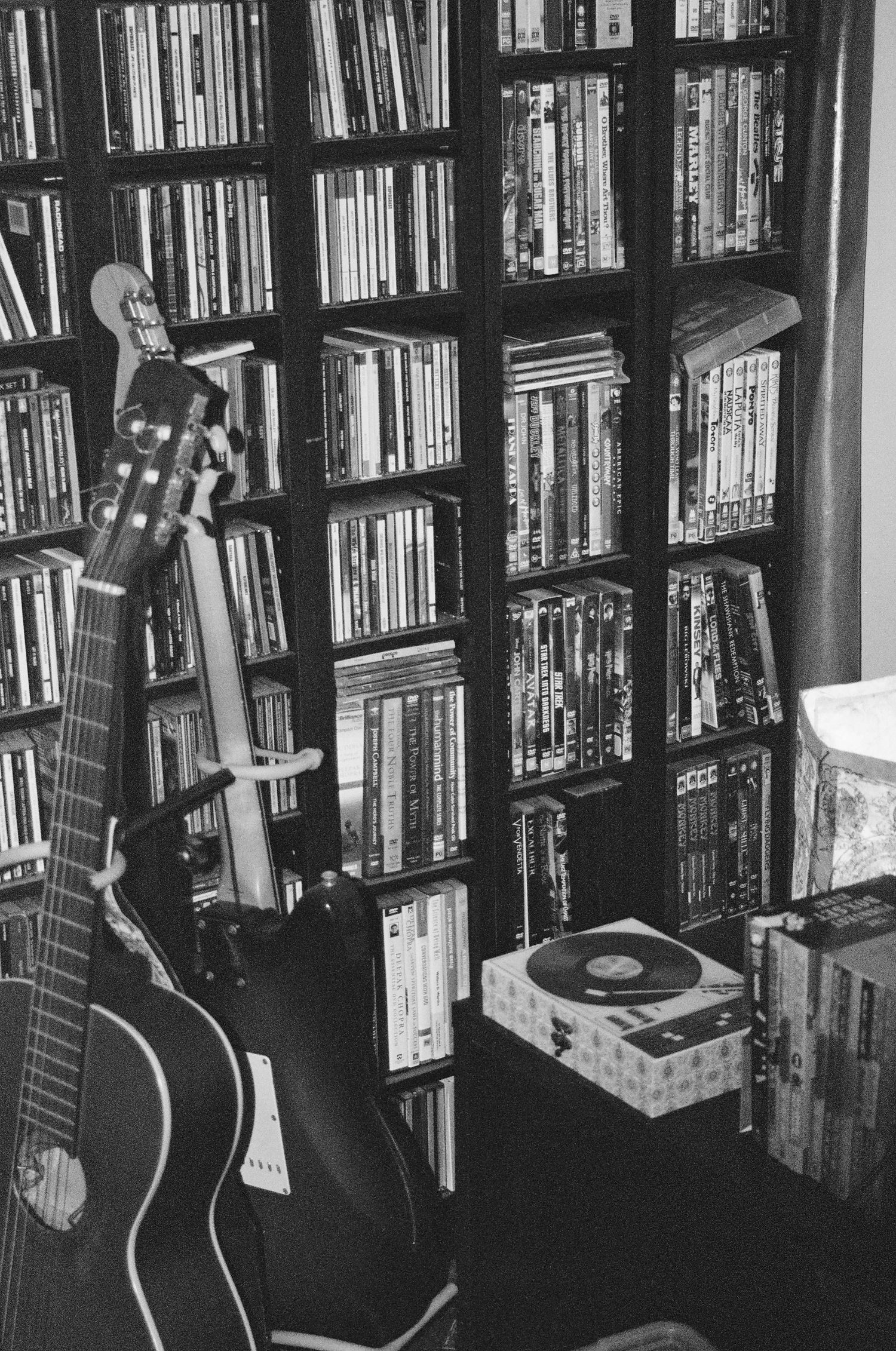 A black and white photo of a guitar next to bookshelves filled with vinyl records and books.