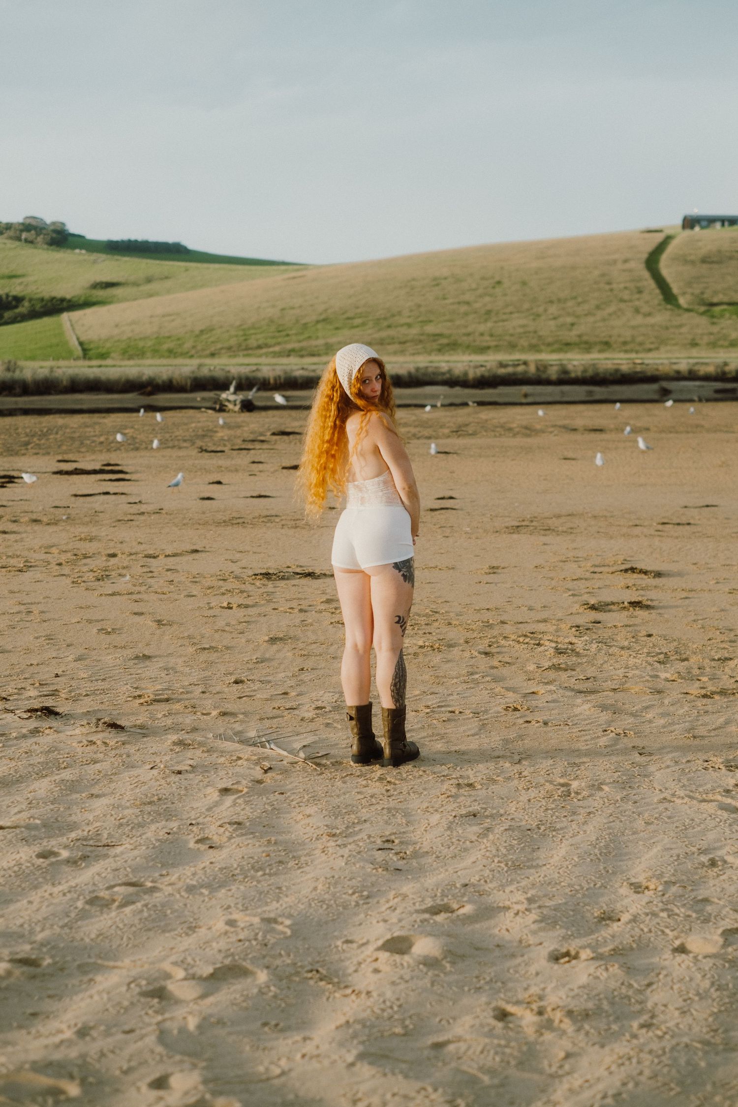 Person with long hair standing on a sandy beach looking at rolling hills in the distance.
