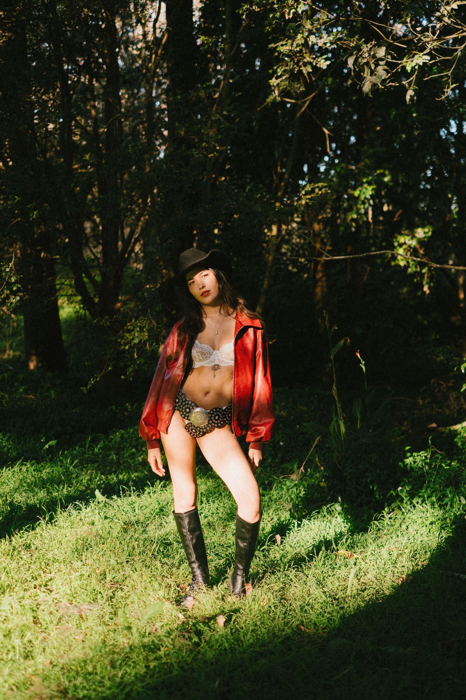 A figure in a red jacket and boots crouches in a sunlit grassy forest clearing.