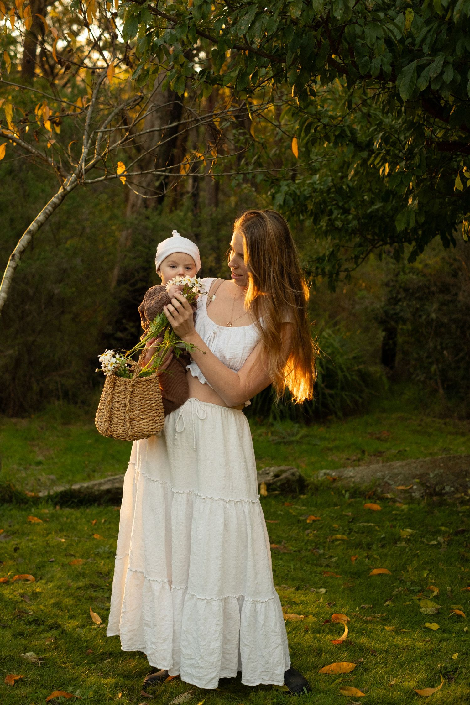 A woman in a white maxi dress holds a baby outdoors in a backlit autumn garden with a basket at sunset.