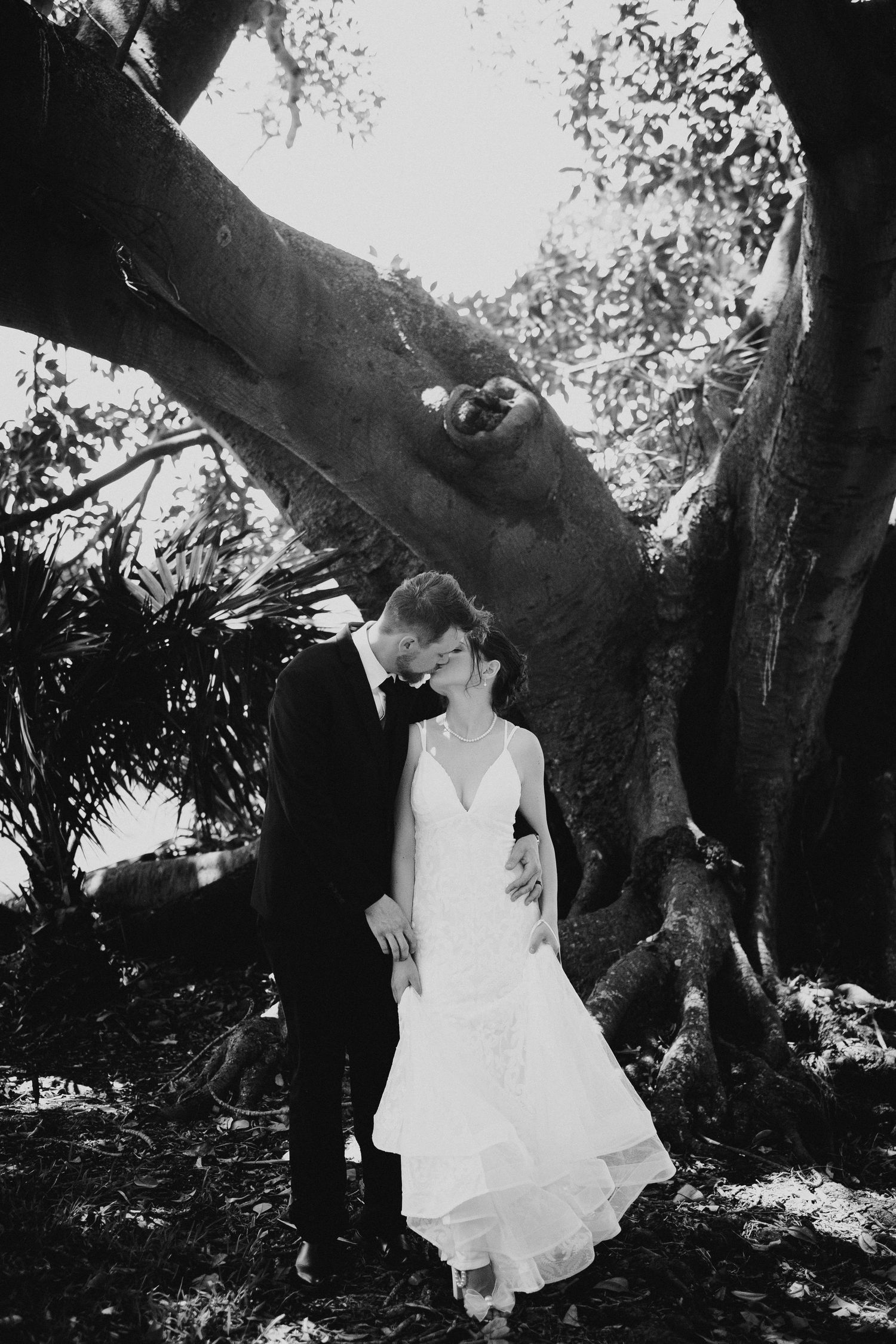 A romantic black and white wedding photo of a couple standing beneath a large tree.