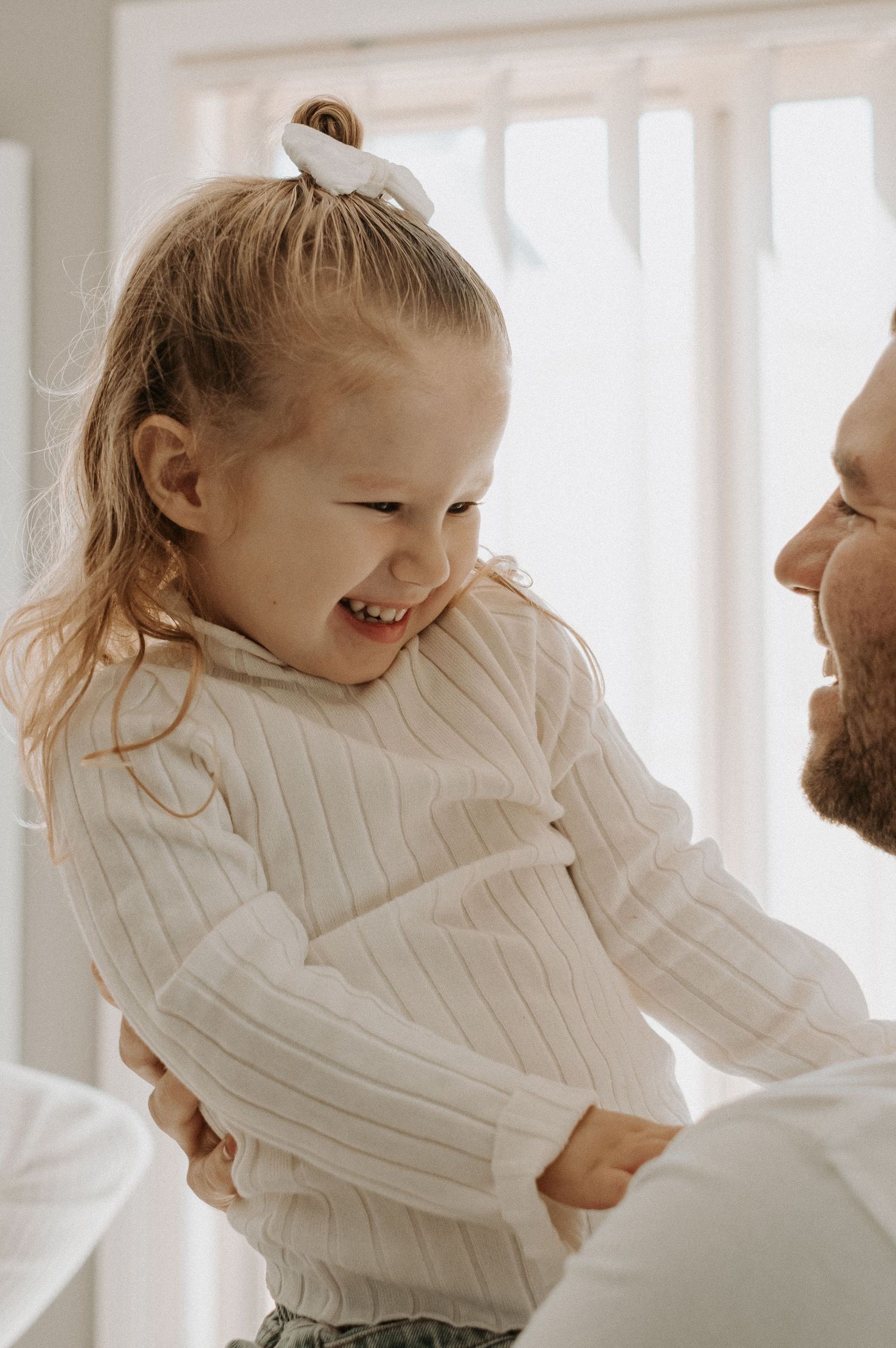 Joyful candid moment shared between parent and child in white sweaters by bright window.