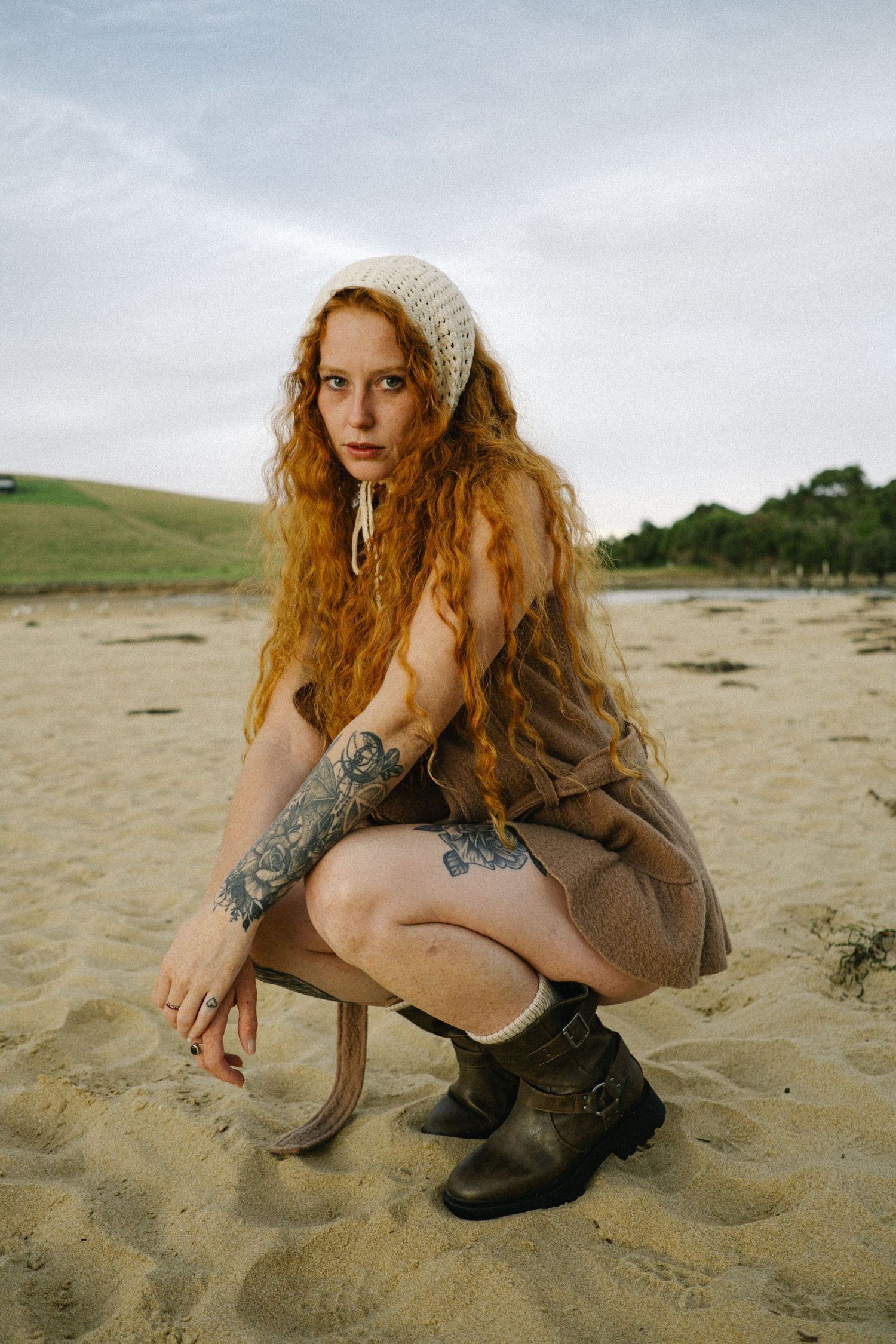 Person in boots and knit hat crouching on sandy beach with rolling hills in background.