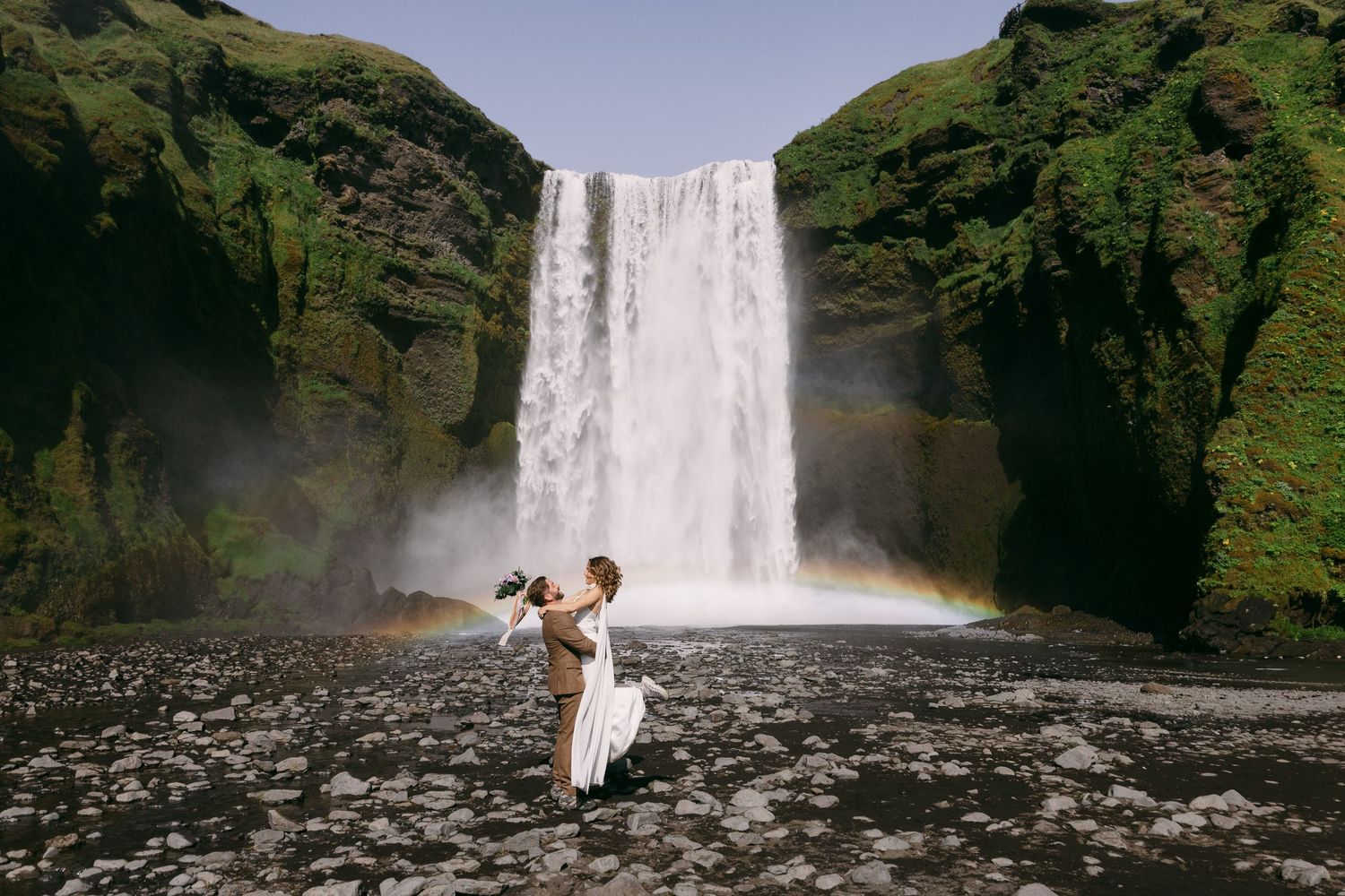 Couple dancing before Skogafoss waterfall in Iceland with a rainbow arching behind them on a misty day.