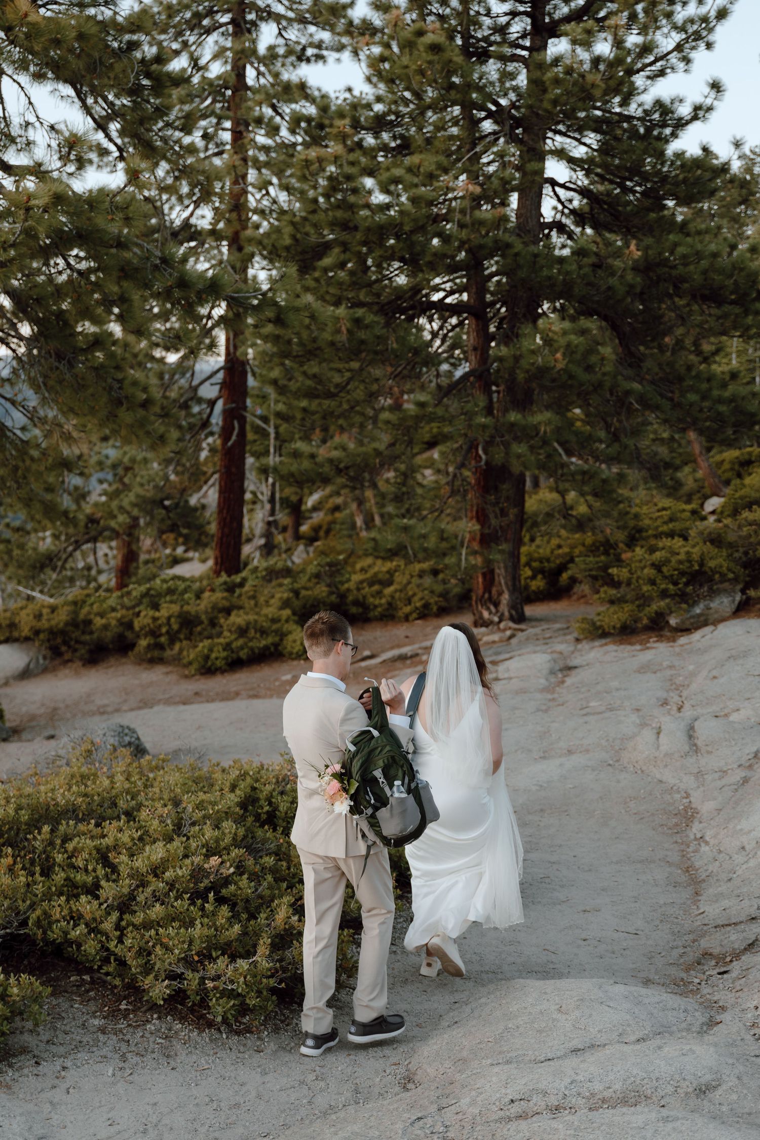 Bride in white gown and veil walks with groom in tan suit on rocky mountain path surrounded by pine trees.