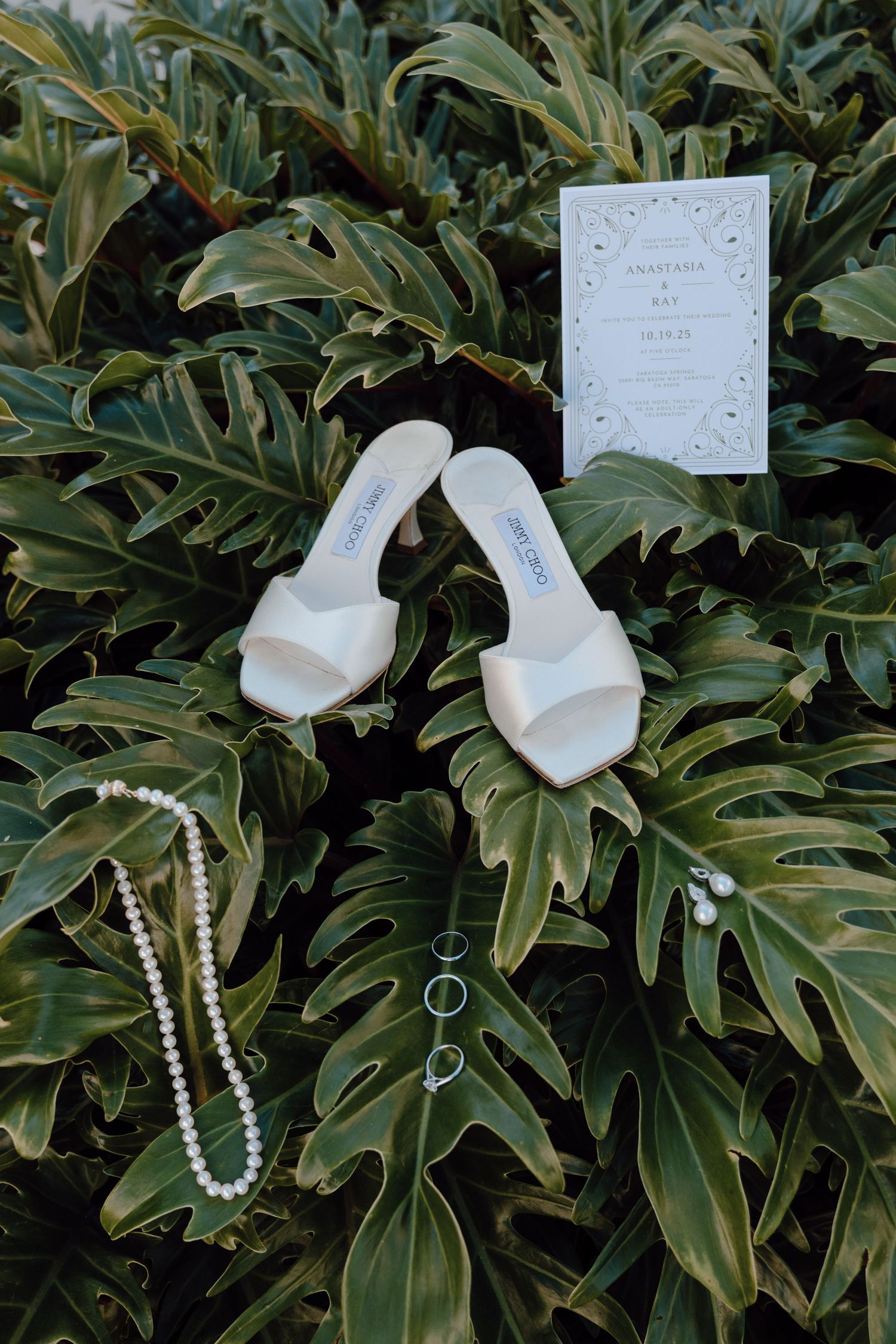 White bridal shoes, pearl necklace and wedding invitation displayed artistically on tropical green monstera leaves.