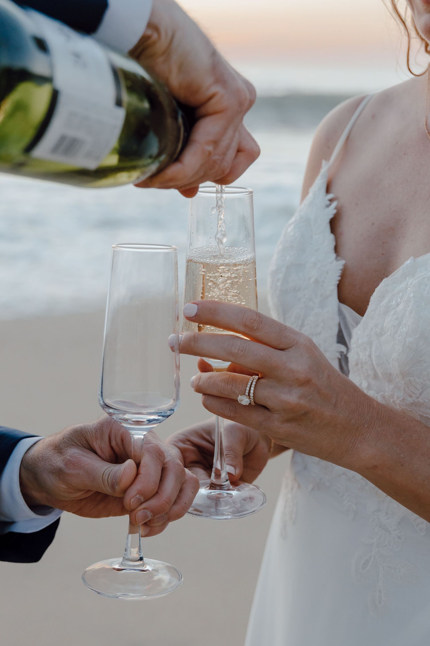 Champagne being poured into flute glasses held by bride and groom on beach at sunset wedding celebration.