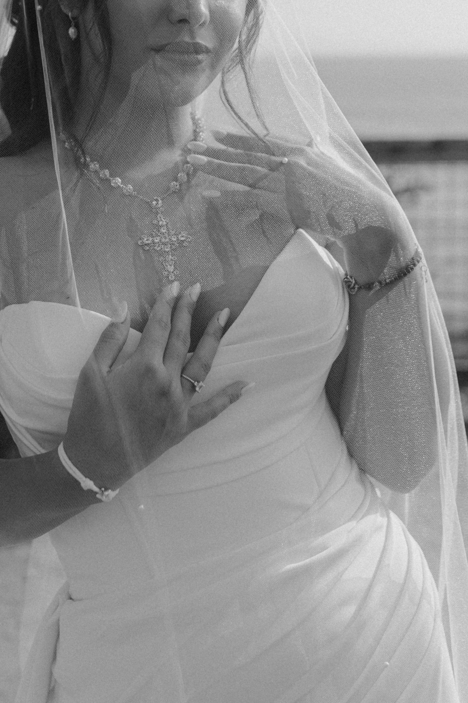 Black and white close-up of a bride wearing an elegant necklace and flowing veil with her hands clasped to her chest.