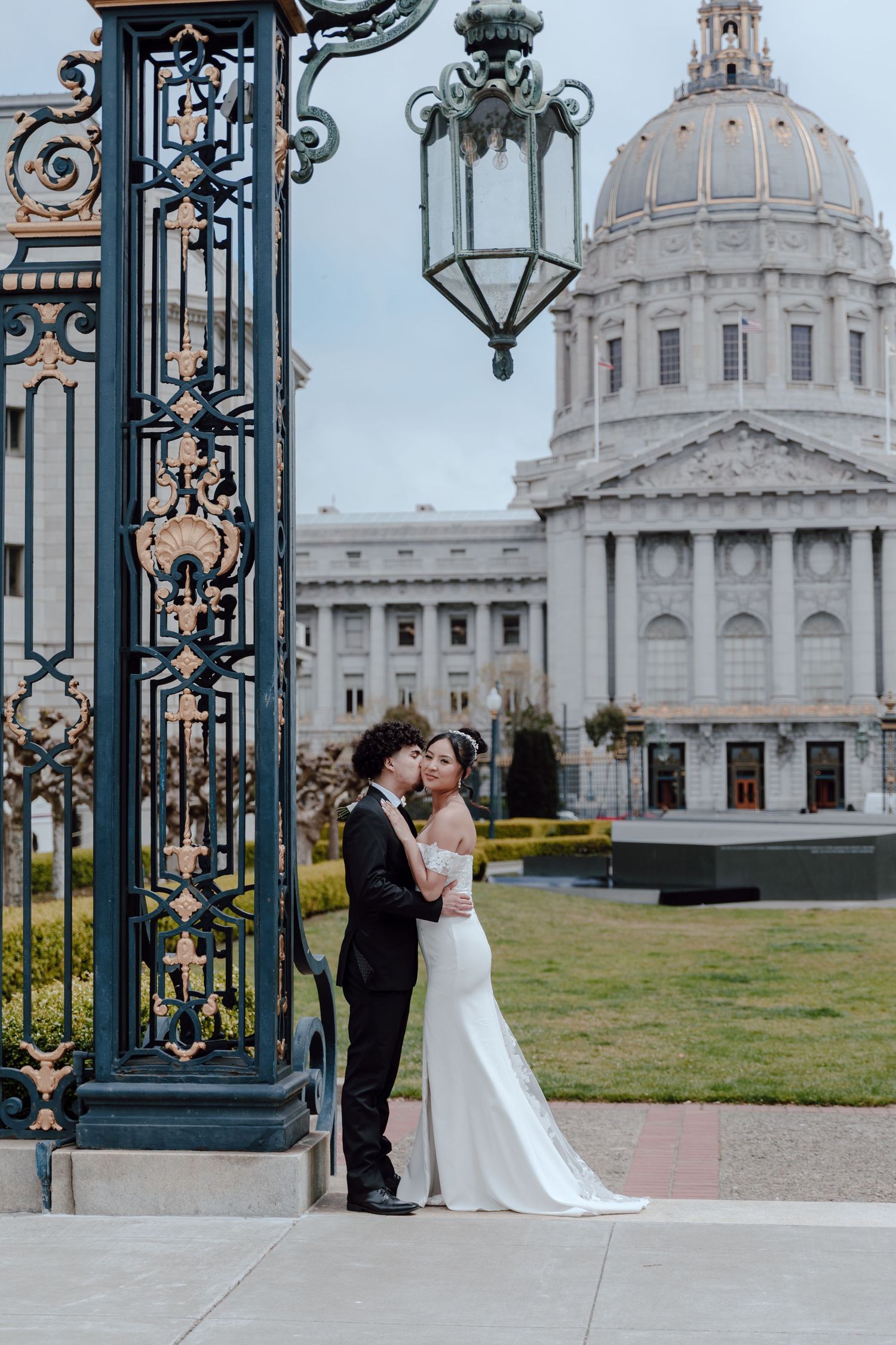 Bride and groom kissing by ornate iron gates with San Francisco City Hall dome in background.