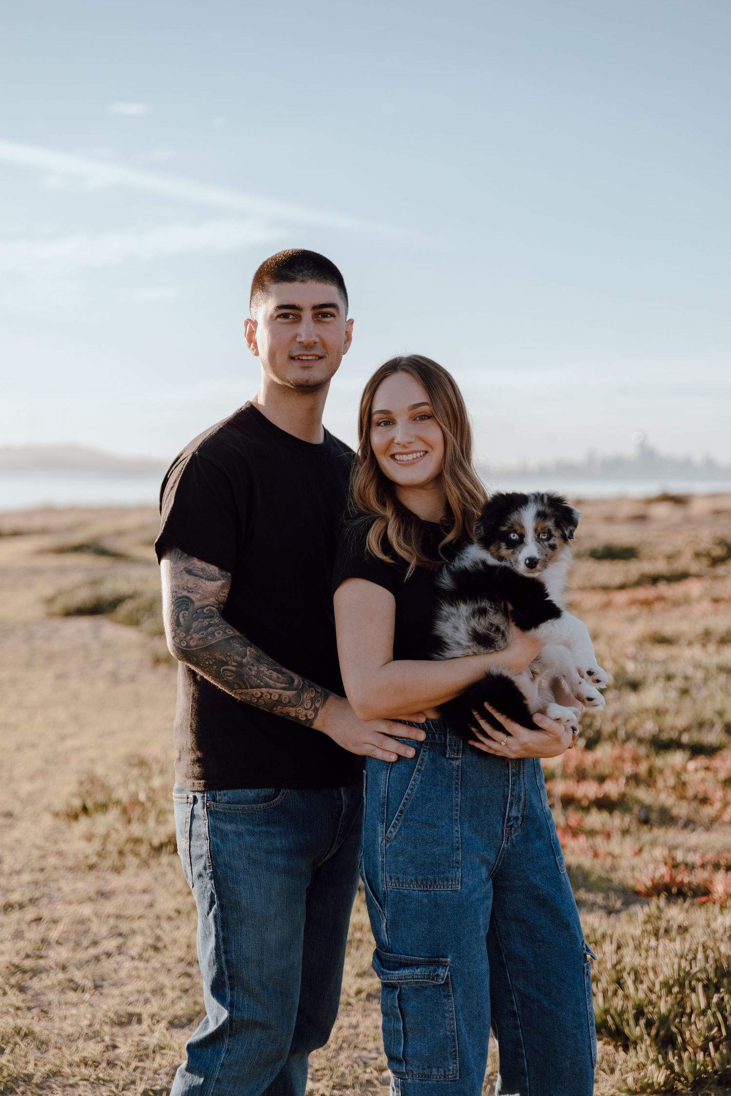 A couple in casual denim outfits poses together with their Australian Shepherd dog against a desert landscape backdrop.