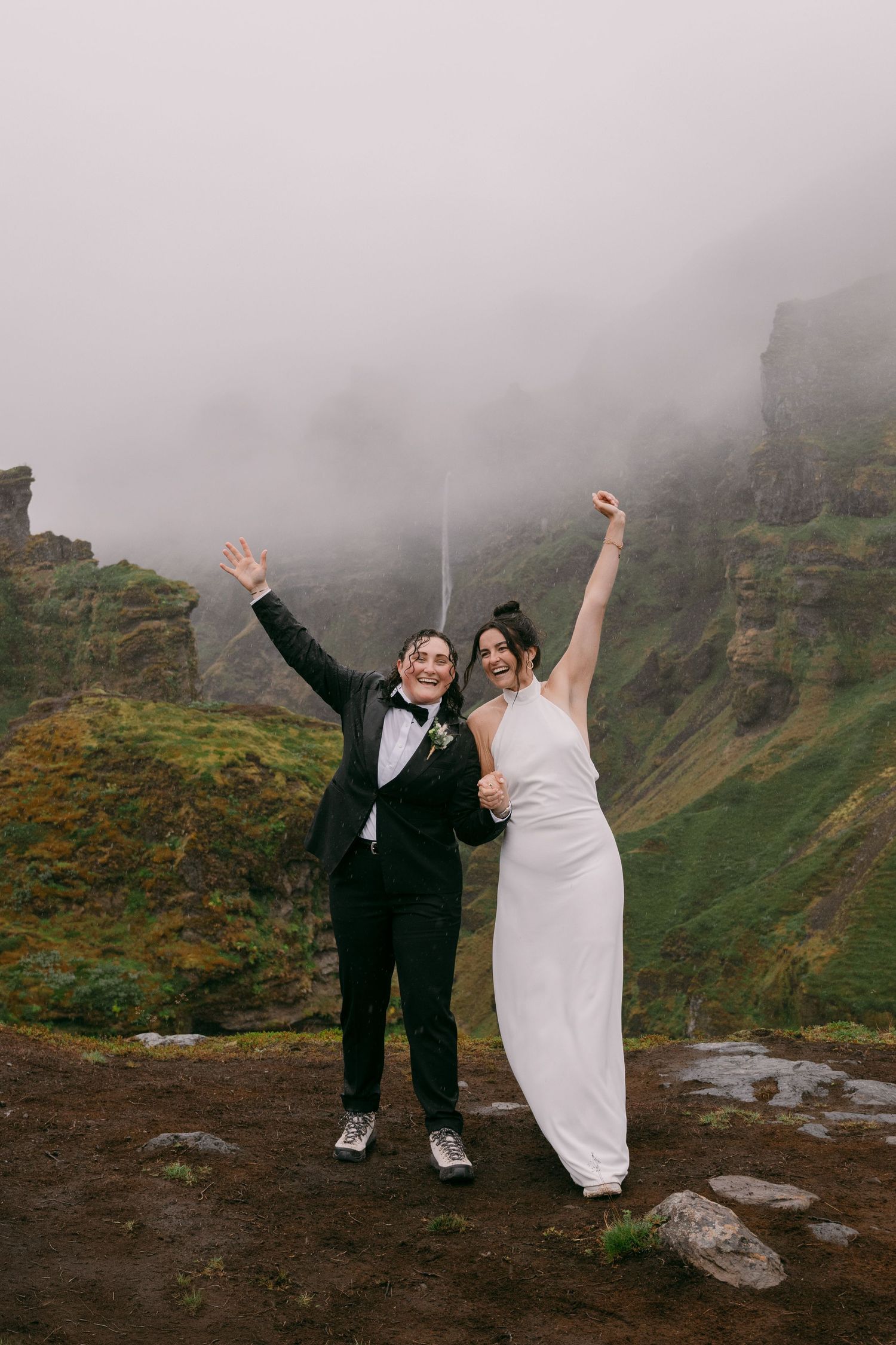 Two brides celebrate joyfully in wedding attire against misty Iceland waterfall cliffs.