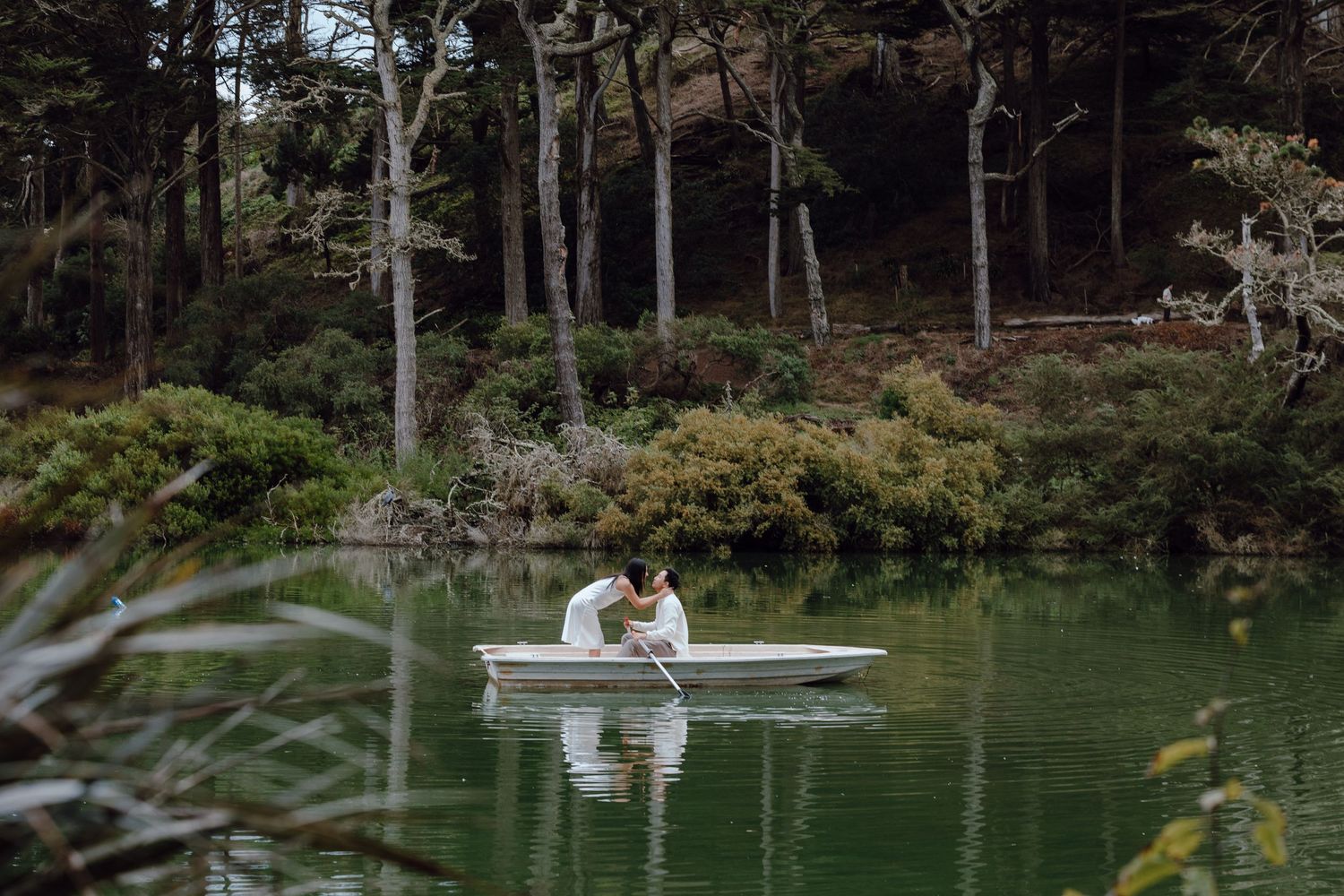 Couple sharing a romantic kiss on a white rowboat on a serene green lake surrounded by lush forest trees.