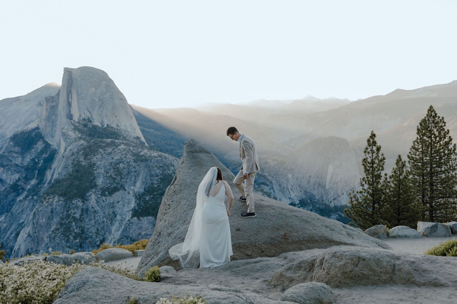 Bride and groom stand on rocky terrain at Glacier Point, Yosemite, with Half Dome and misty valley views behind them.
