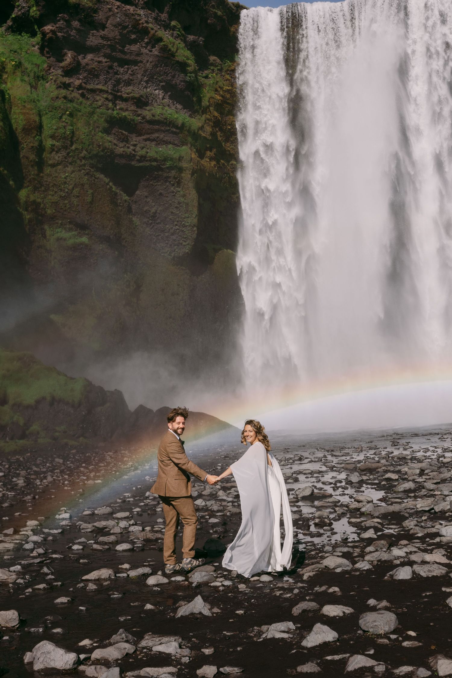 Couple holds hands before Skogafoss waterfall in Iceland with a rainbow arching behind them on their wedding day.