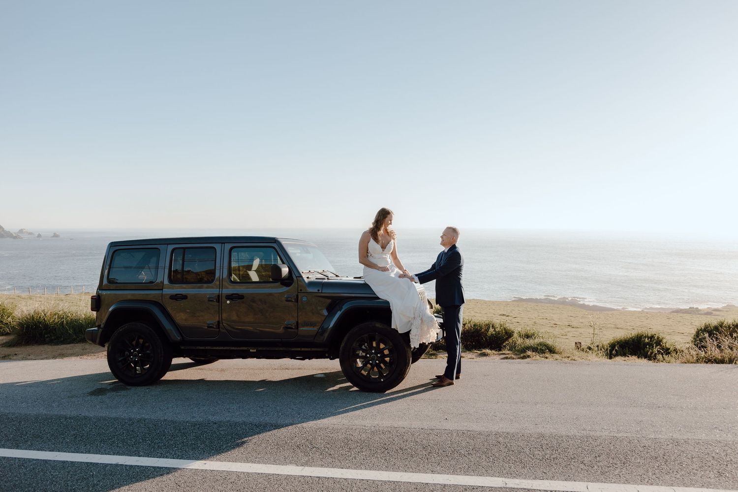 Bride in white gown sits on Jeep hood as groom holds her hand, with ocean and clear sky backdrop.