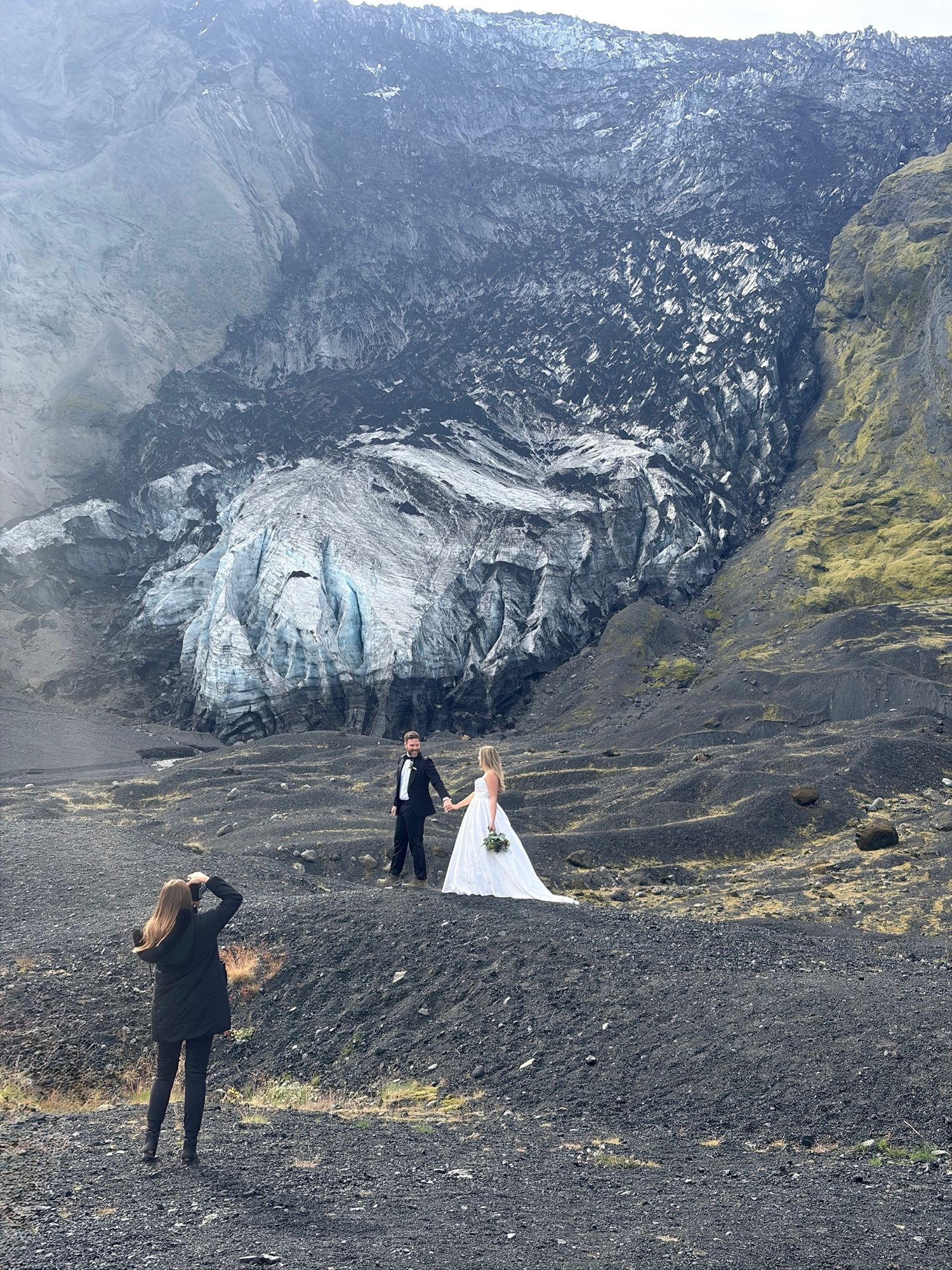 Couple in wedding attire posing for photos in front of a glacier on a volcanic black sand landscape in Iceland.