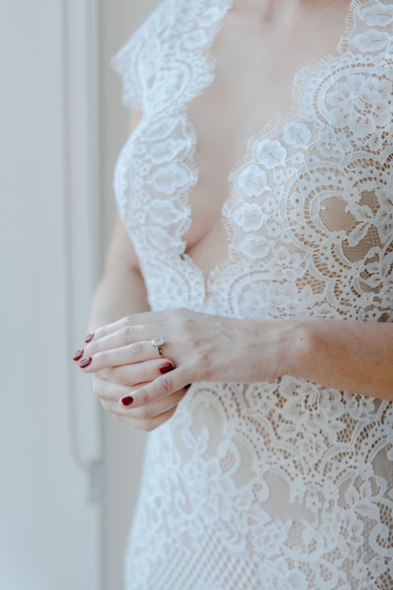 Close-up detail of delicate white lace wedding dress with hands showing red nail polish and engagement ring.