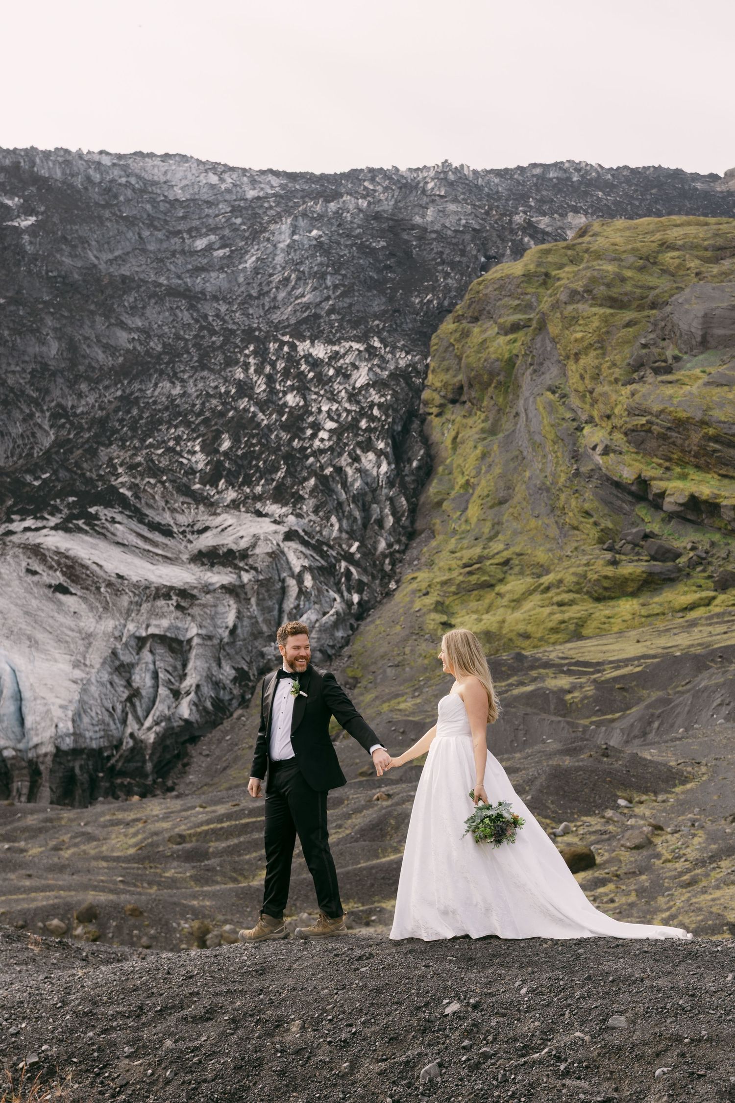 Bride and groom hold hands before a dramatic glacier and green volcanic hillside in Iceland wedding photo.