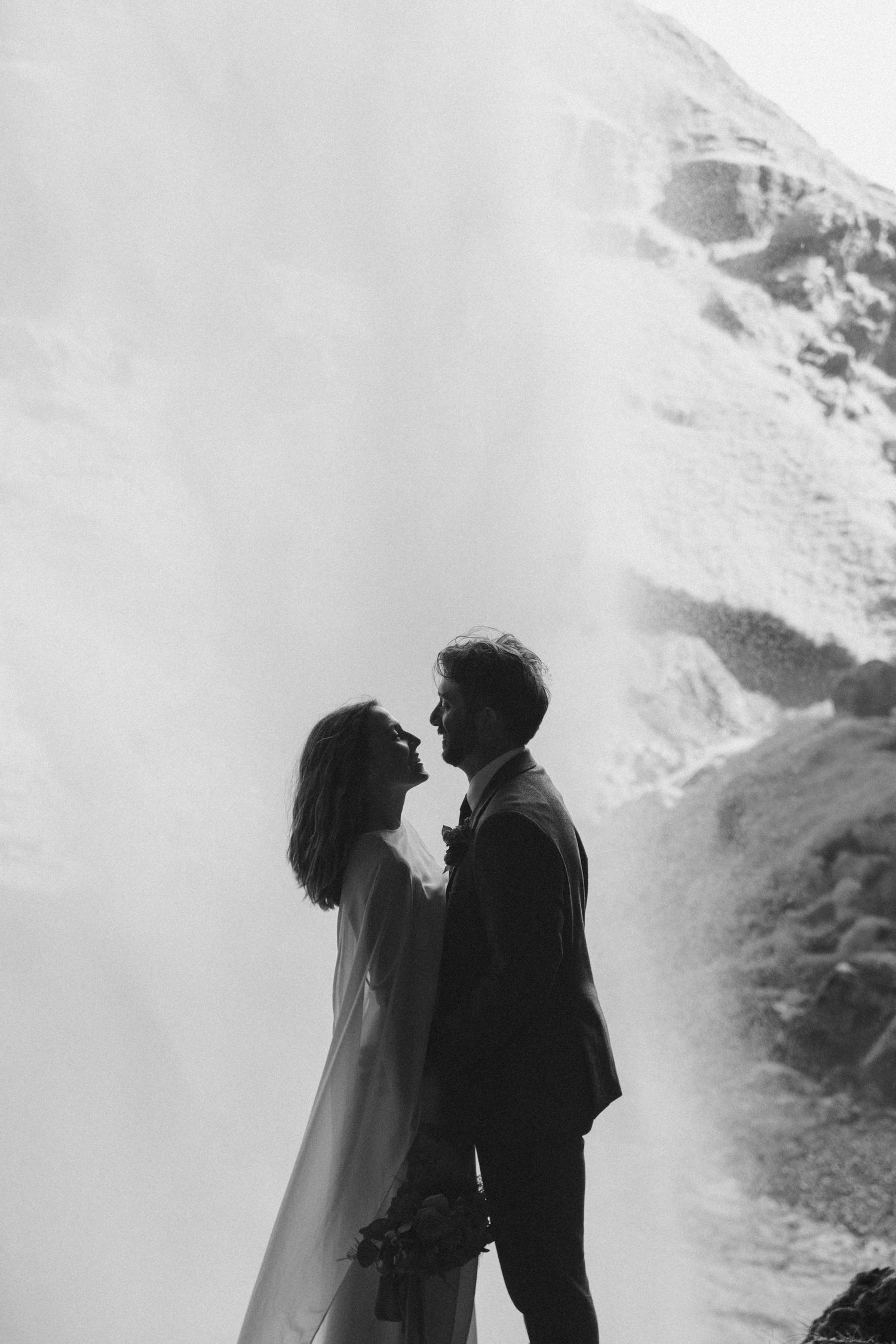 Black and white photo of a couple in wedding attire sharing a romantic moment in front of a snowy mountain waterfall.