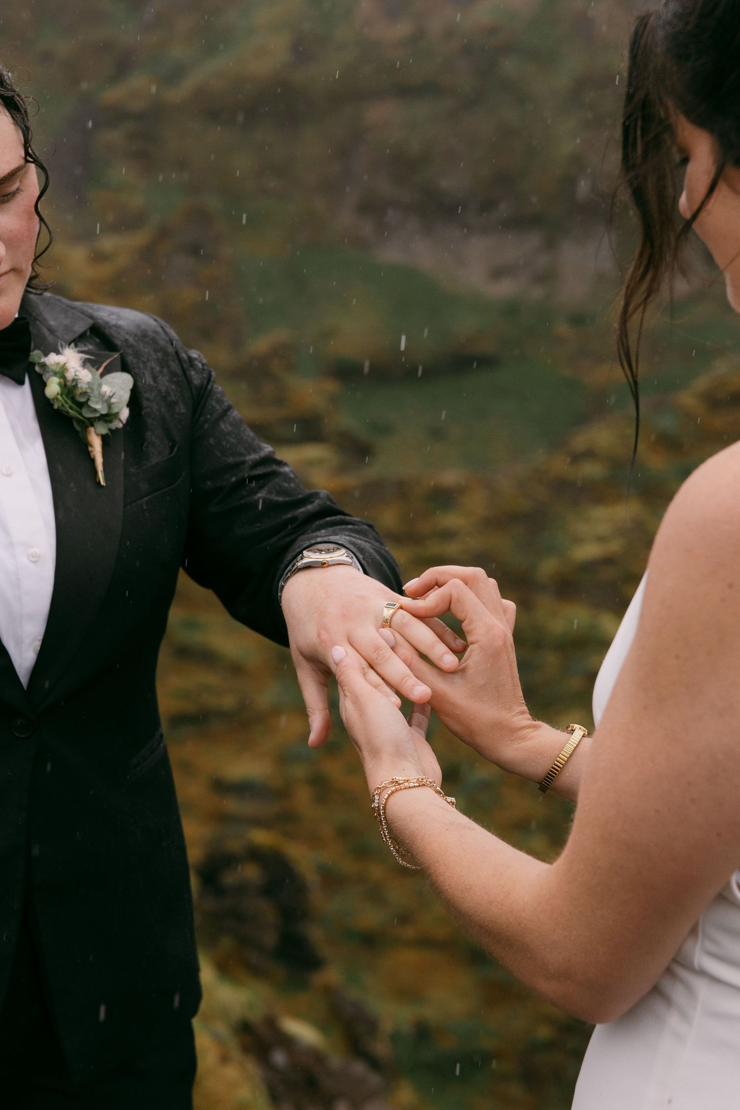 Two brides exchanging wedding rings outdoors in the rain, one wearing a dark suit with boutonniere, the other in a white dress.