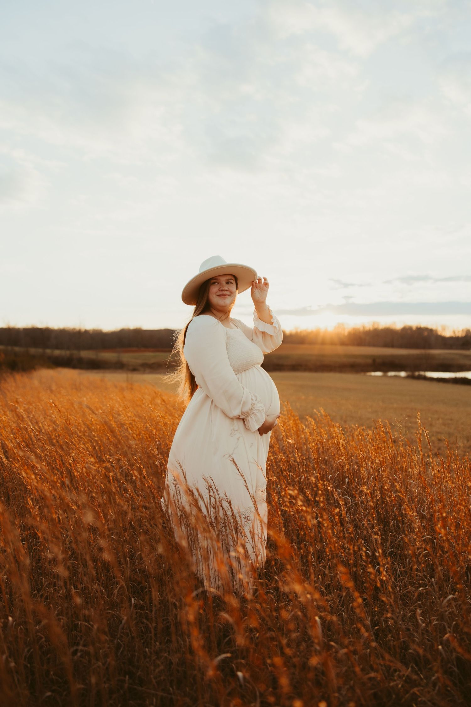 Pregnant woman in white dress and hat standing in golden field at sunset during maternity session.