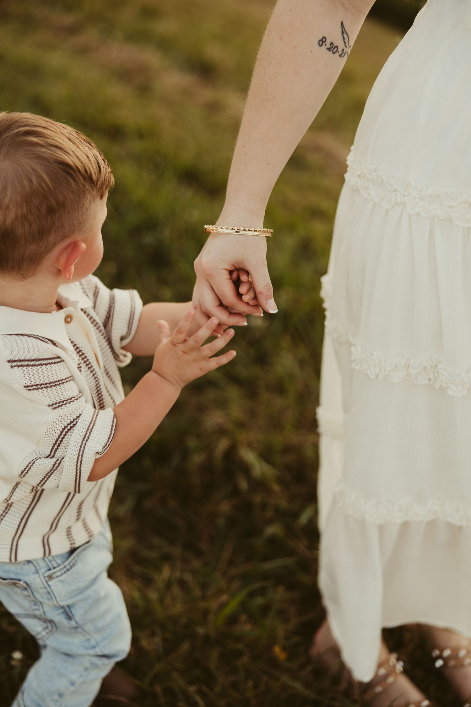 Young toddler boy holding his mothers hand while walking through a green grassy field at sunset in Ruffin, NC.