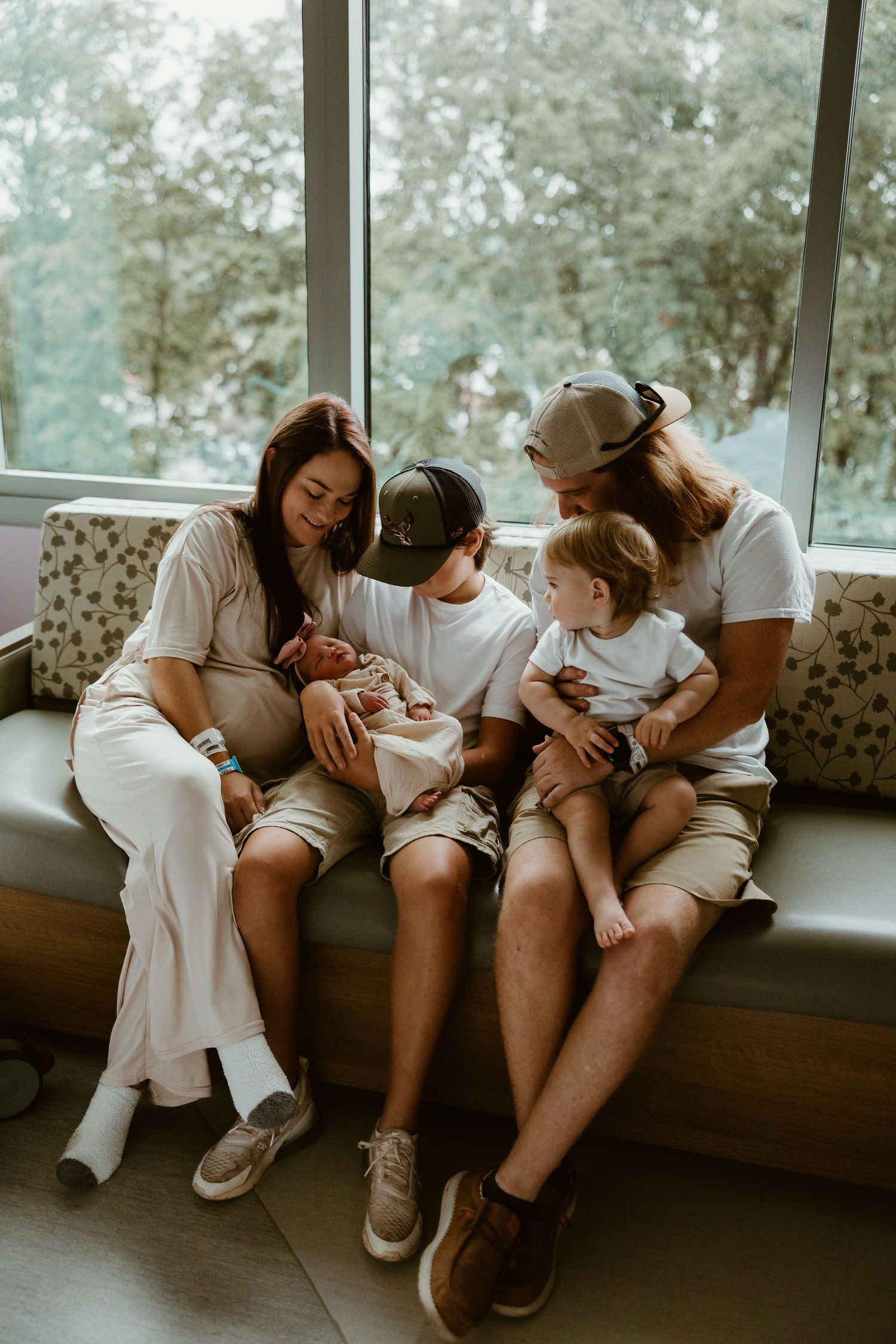 A family of five sits by a large window, lovingly gazing at their newborn baby in a hospital room at Women's Hospital in Greensboro, NC.