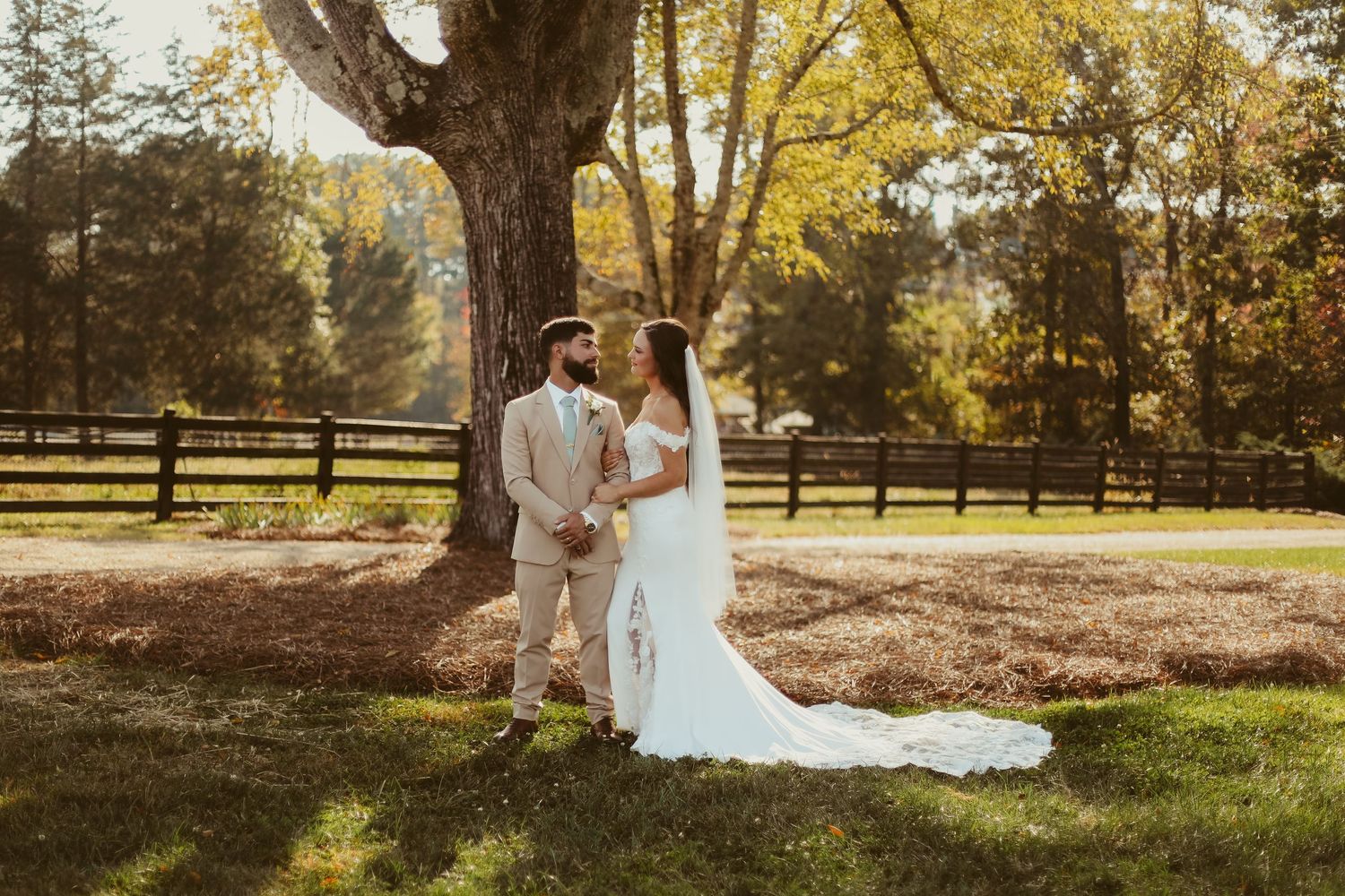 Bride and groom hold hands under a large oak tree at a sunny outdoor farm wedding venue.