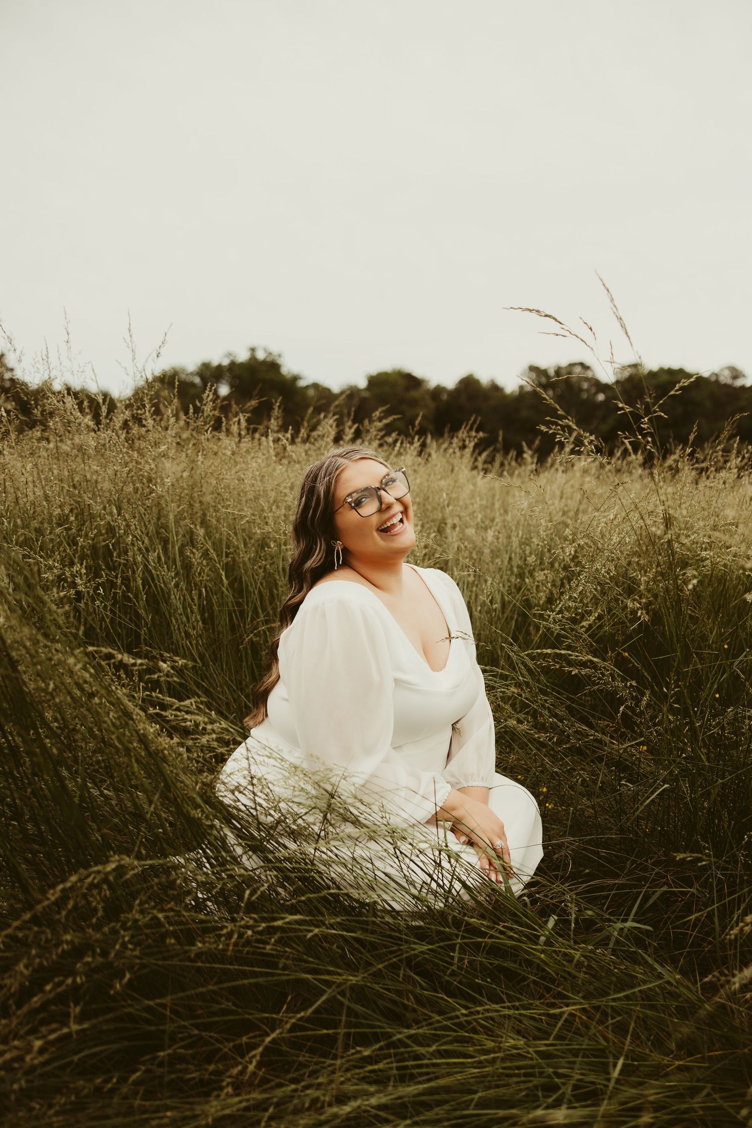 A smiling woman in a white dress sits in a tall green grass field, wearing sunglasses on an overcast day.