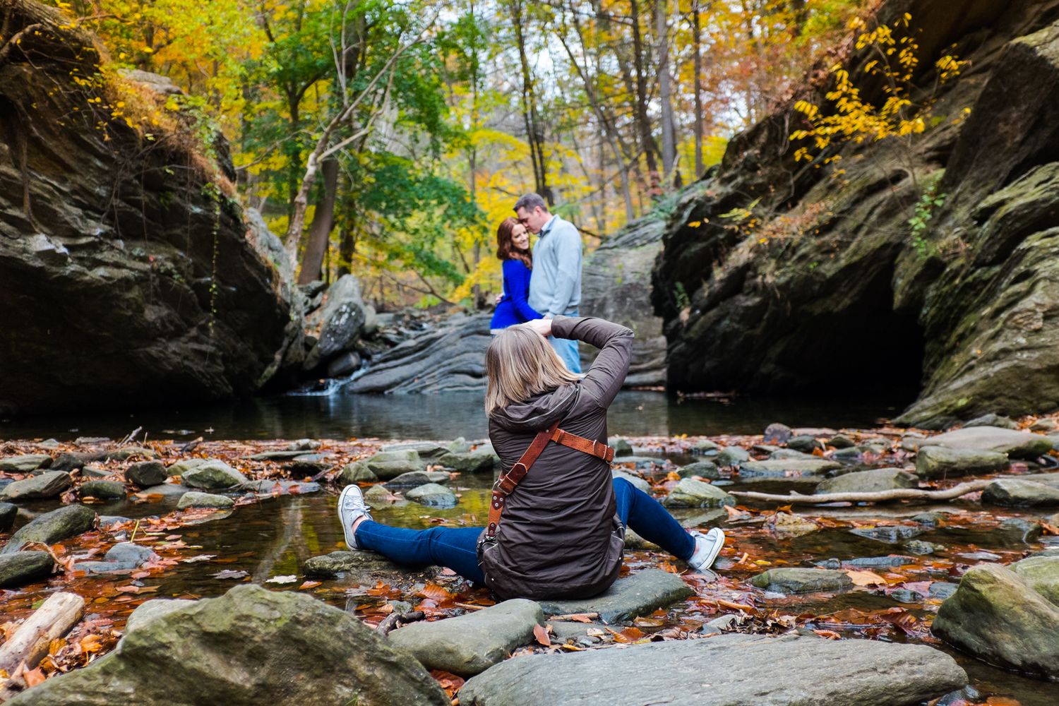 A photographer sits on rocky terrain photographing a couple in blue against autumn foliage in a scenic gorge.