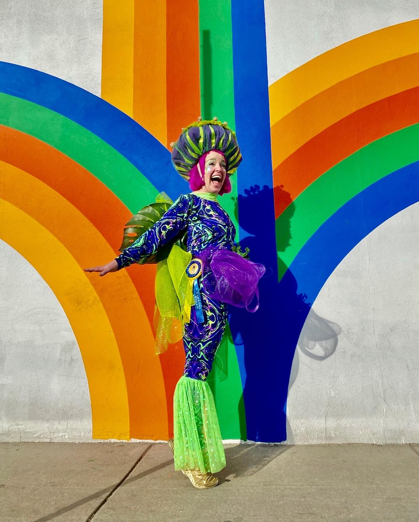 Performer in colorful costume poses dramatically against rainbow wall mural.