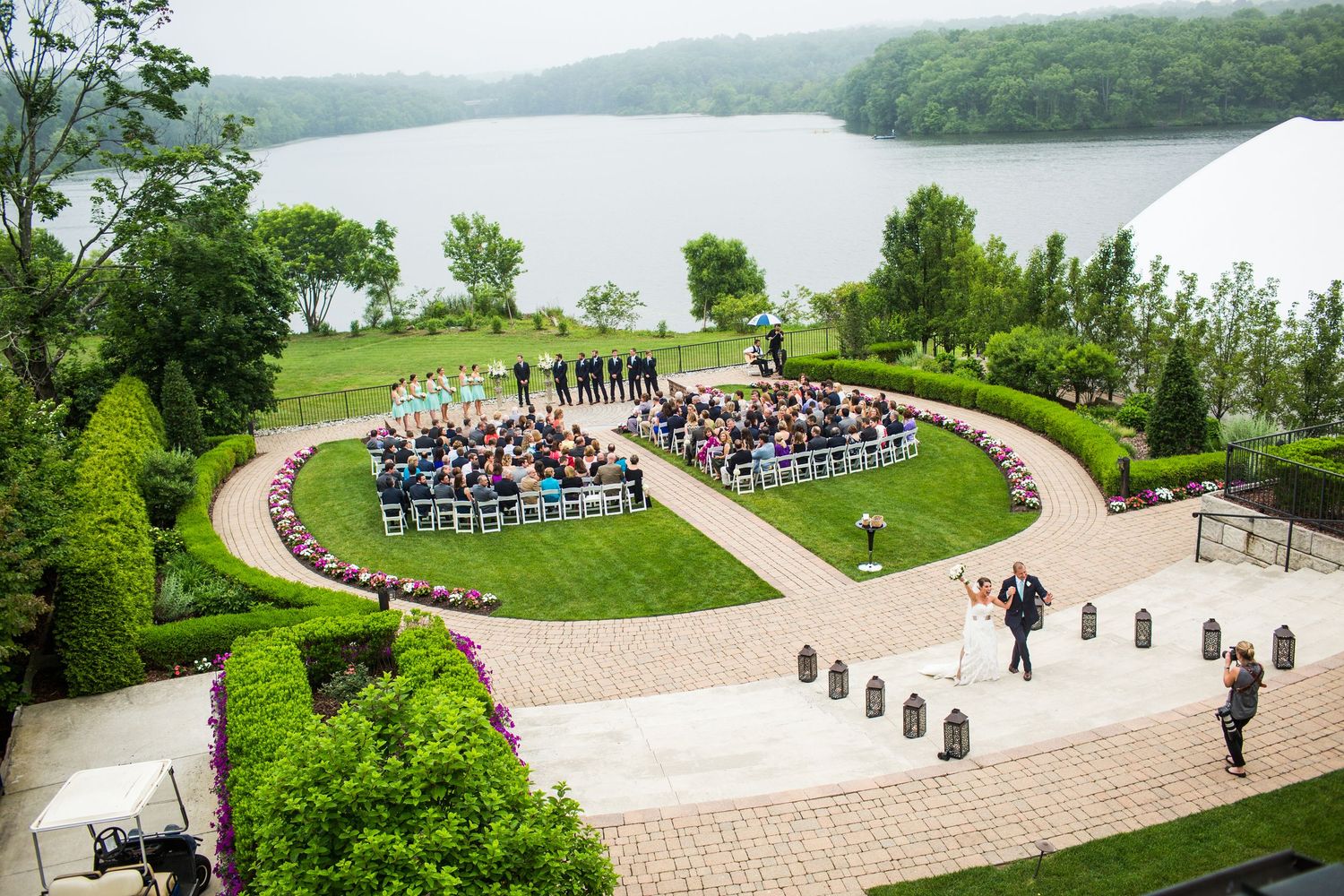 Aerial view of an elegant outdoor wedding ceremony in a circular garden overlooking a serene lake.