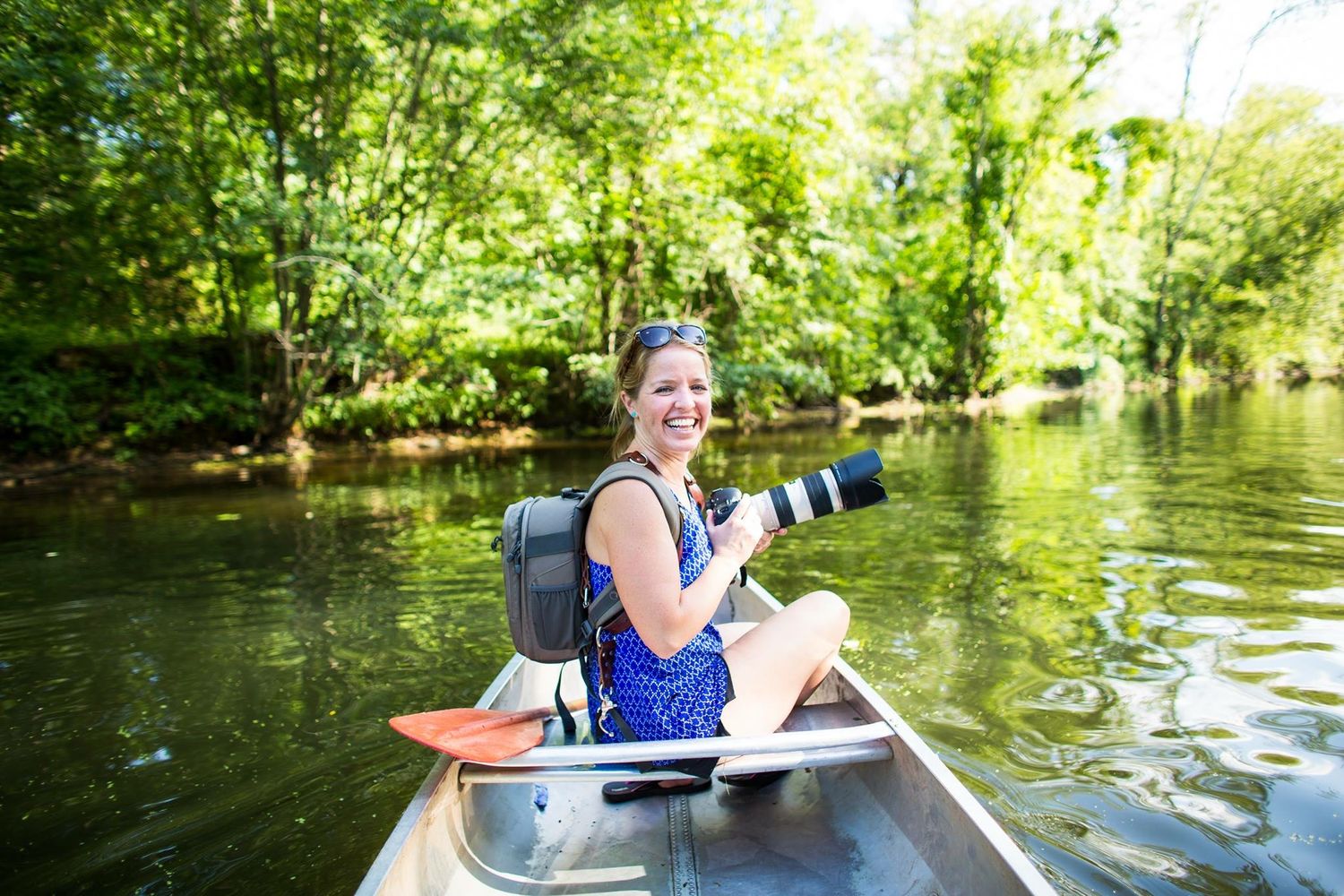 Photographer with camera gear sits in canoe on peaceful river surrounded by green trees.