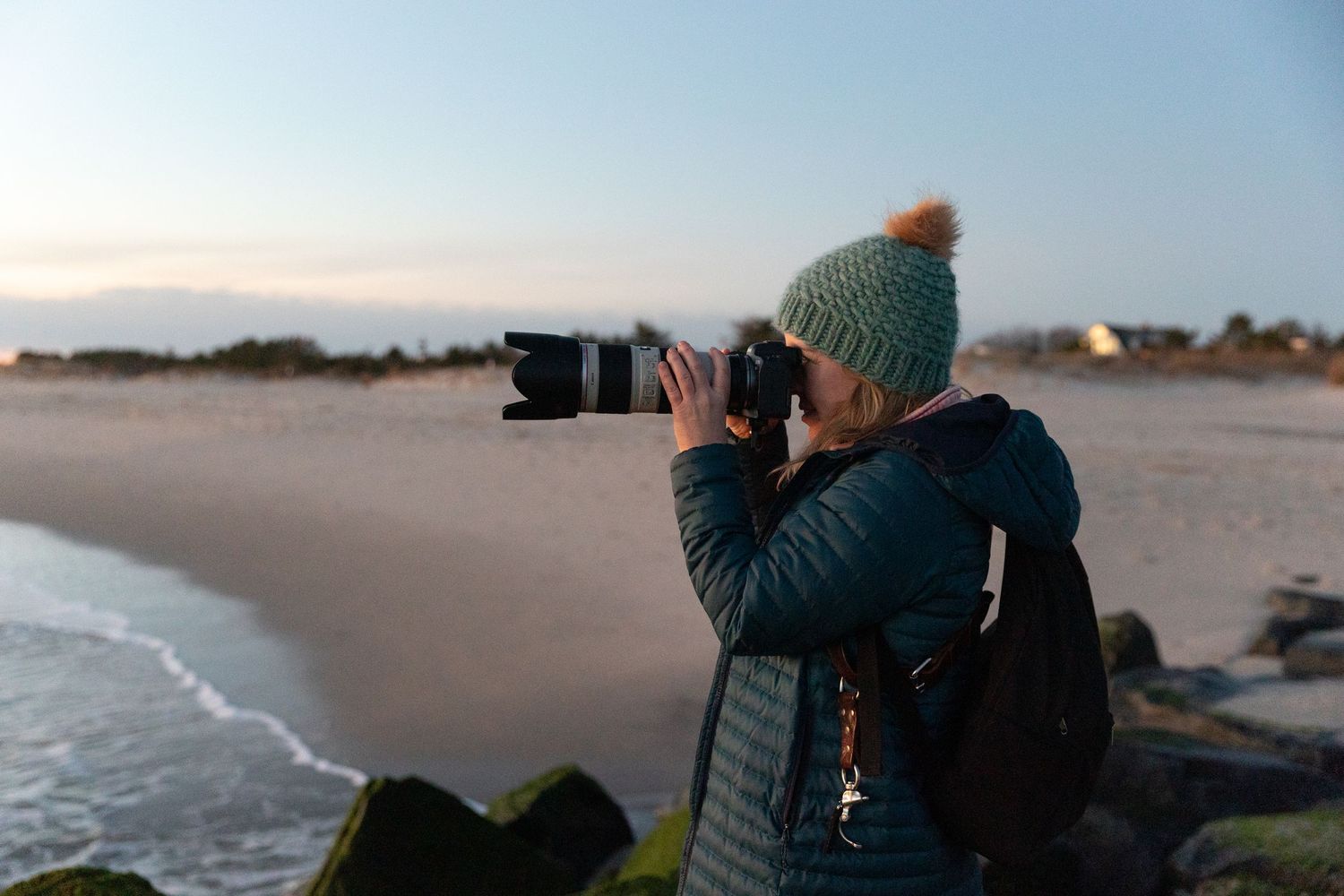 A photographer wearing a teal beanie captures sunset photos at a beach.