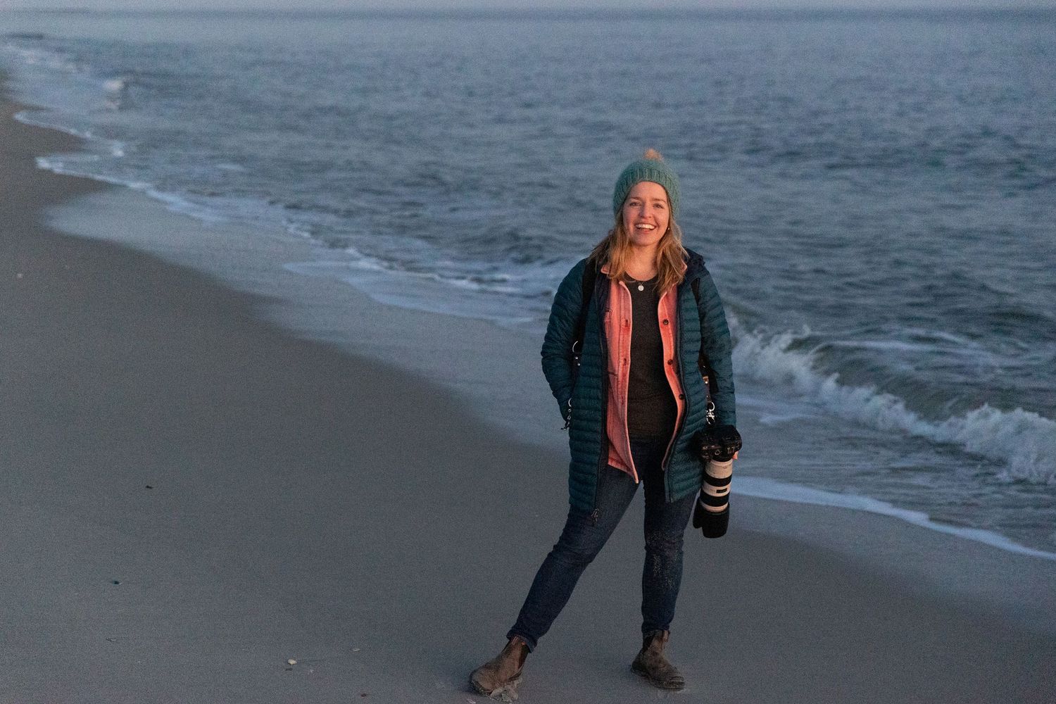 A photographer stands on a beach at dusk holding professional camera equipment.