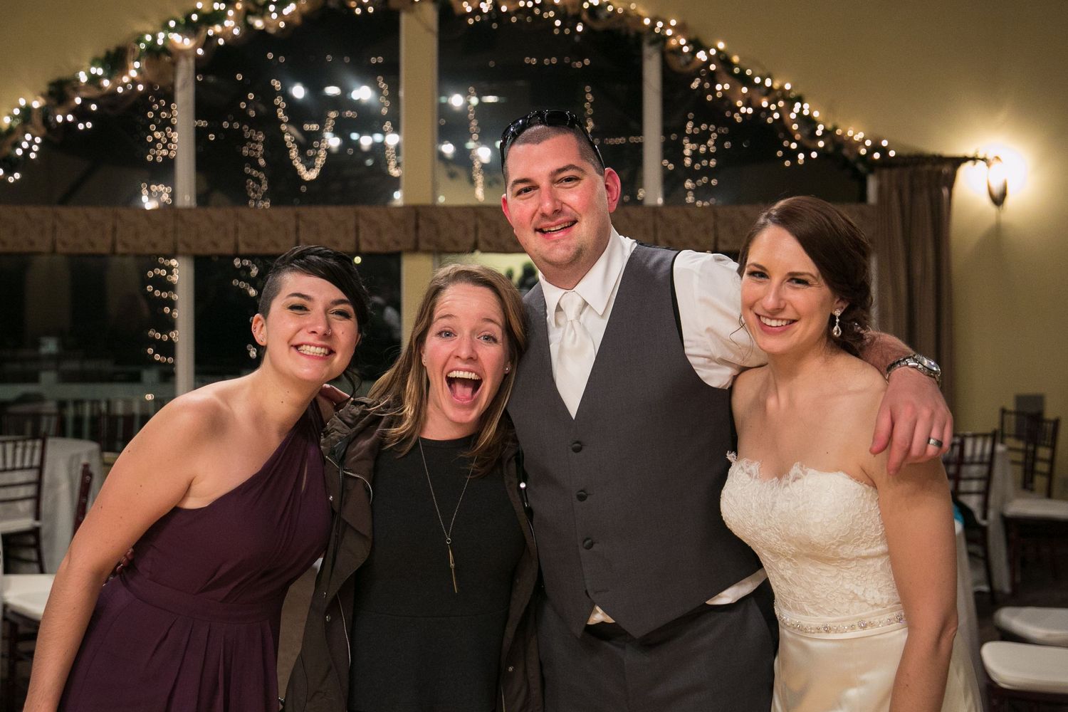 Wedding celebration photo showing group of four people smiling together under decorative string lights at reception venue.