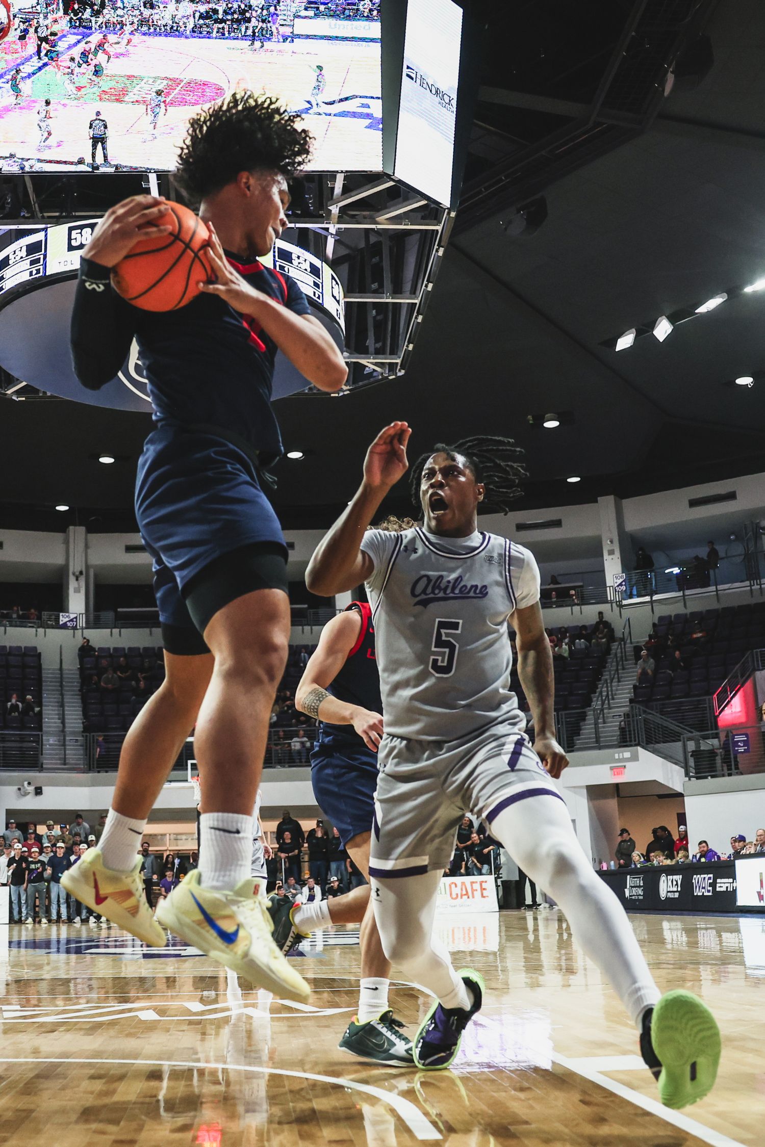 A basketball player in red leaps with the ball as a defender wearing number 5 challenges near the baseline.