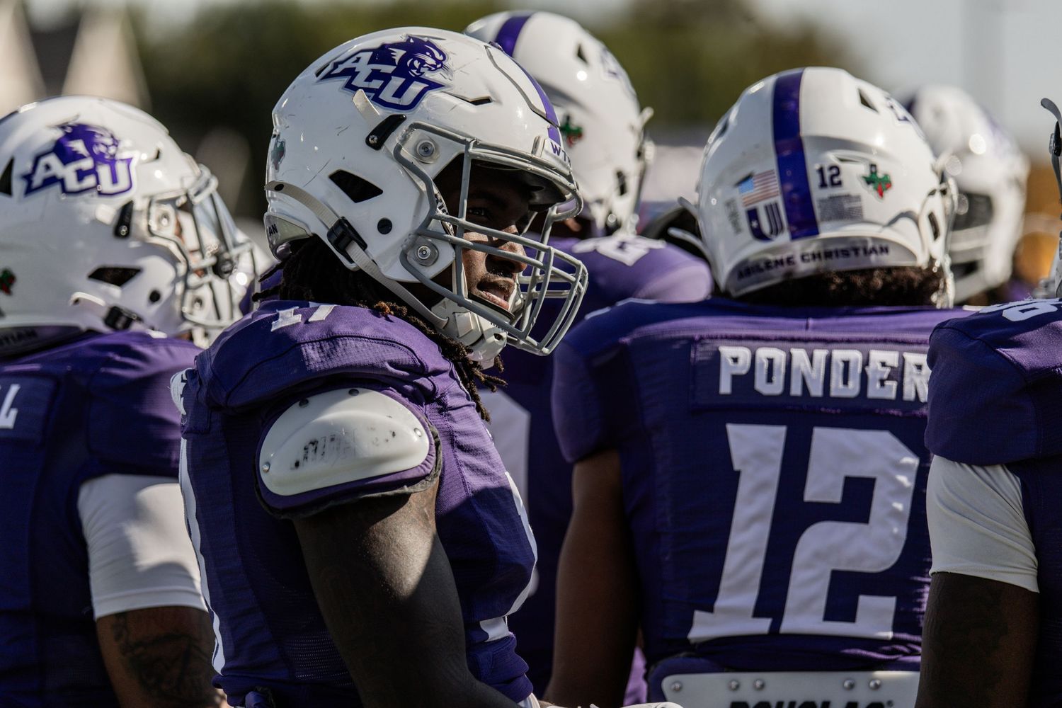 ACU football players in purple and white uniforms huddle together on the field.