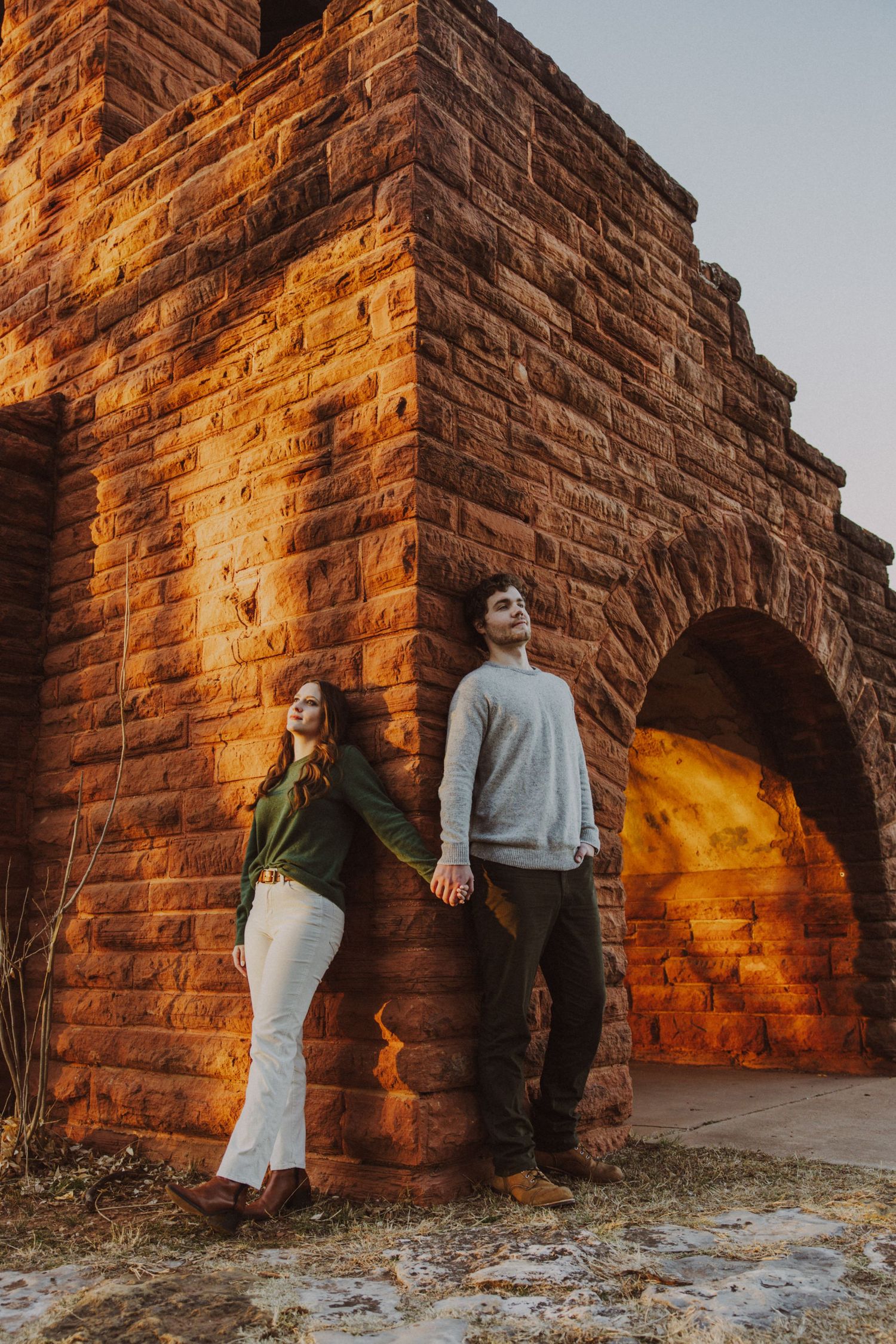 Couple holding hands beside a rustic stone arch structure at sunset in warm golden light.