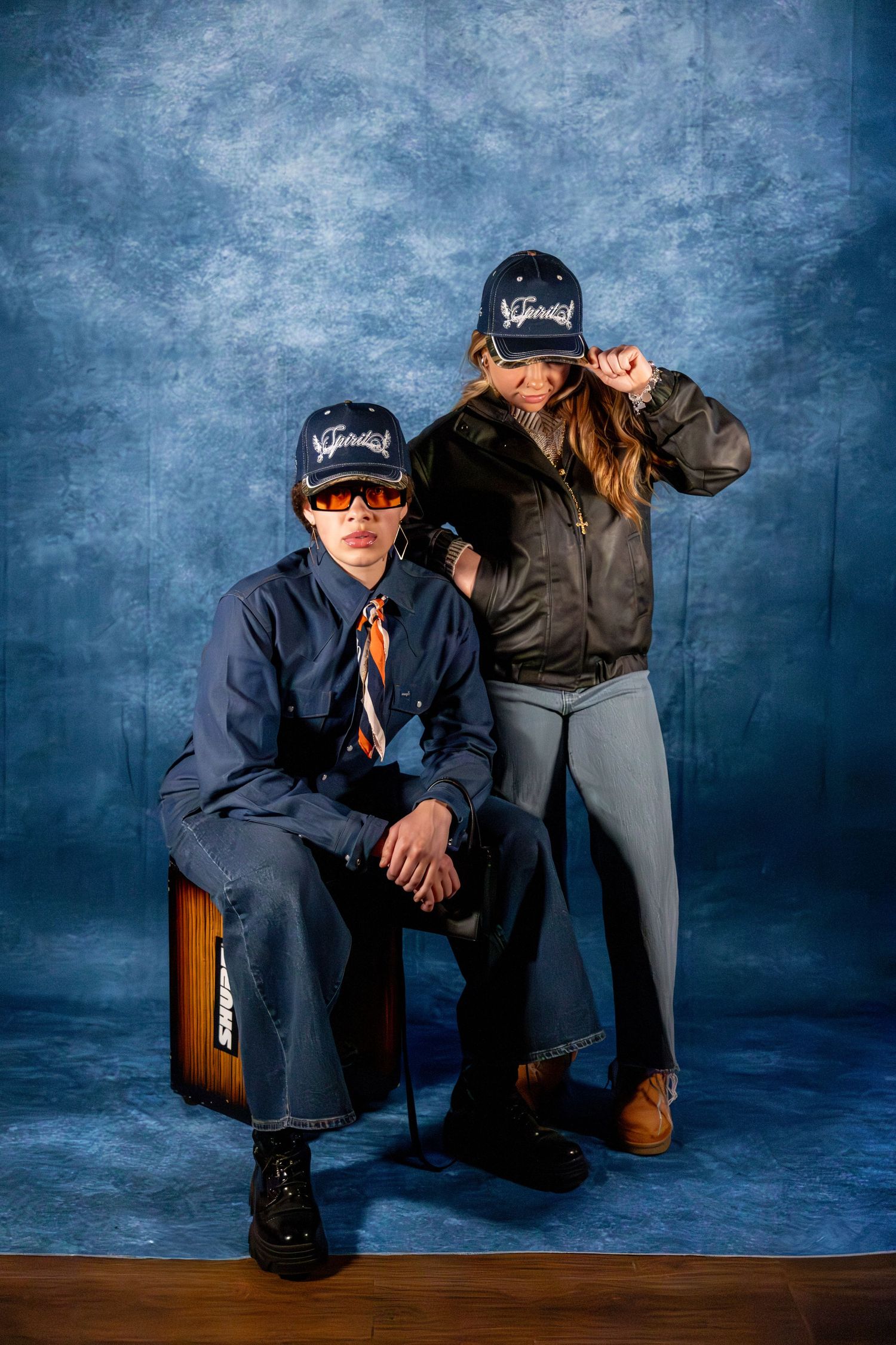 Two models wearing matching navy caps pose together against a blue textured background.